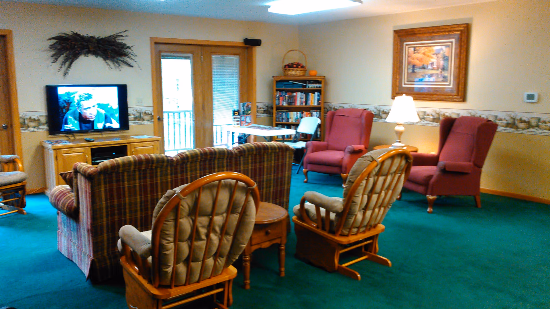 A cozy living room area with green carpet, a plaid sofa, two cushioned wooden rocking chairs, two red armchairs, a wooden side table with a lamp, a bookshelf filled with books, a TV on a wooden stand, and a framed painting on the wall.