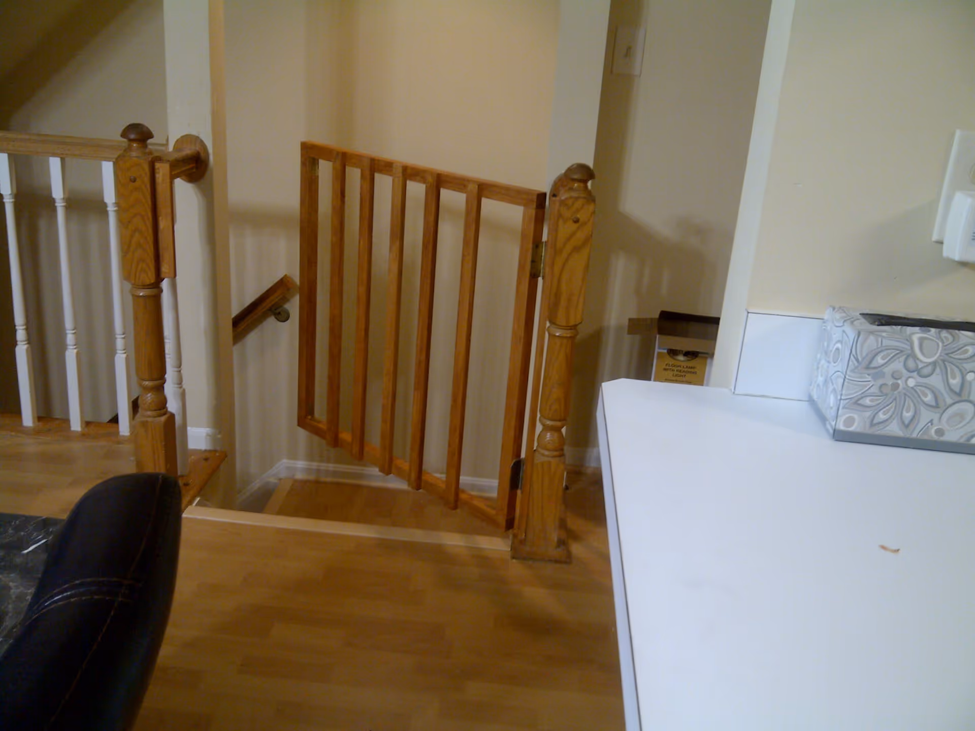 Interior view of a home showing a wooden safety gate at the top of a staircase, wooden handrails, a countertop with a decorative tissue box, and part of a black chair.
