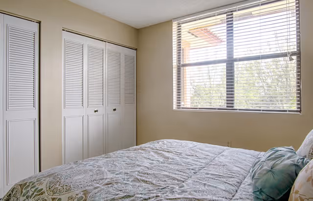A bedroom with a bed covered in a patterned bedspread and several pillows. There is a large window with blinds letting in natural light, and white louvered closet doors along one wall. The walls are painted a light beige color.