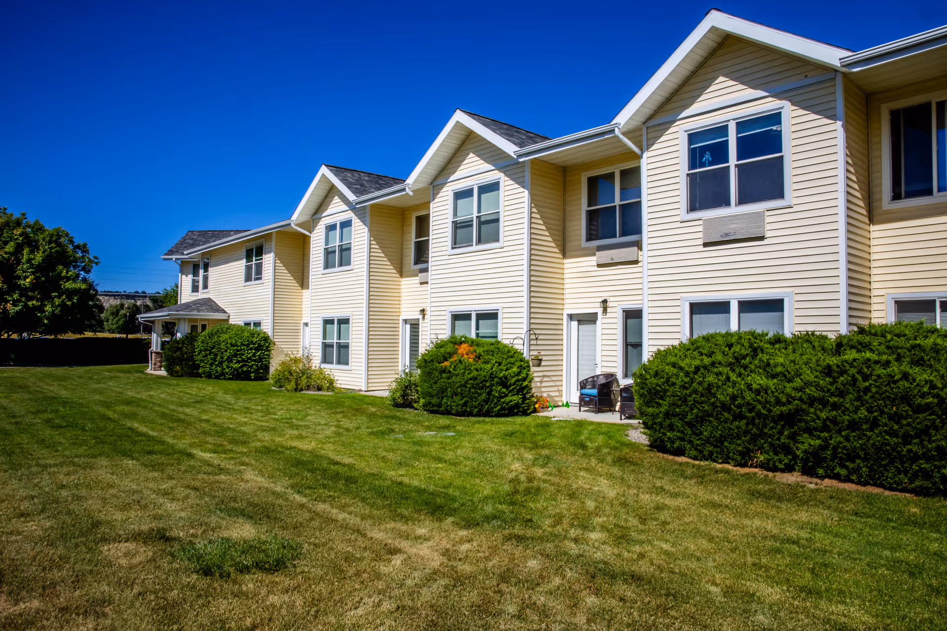 Exterior view of a two-story beige residential building with multiple windows and doors, surrounded by green bushes and a well-maintained lawn under a clear blue sky.
