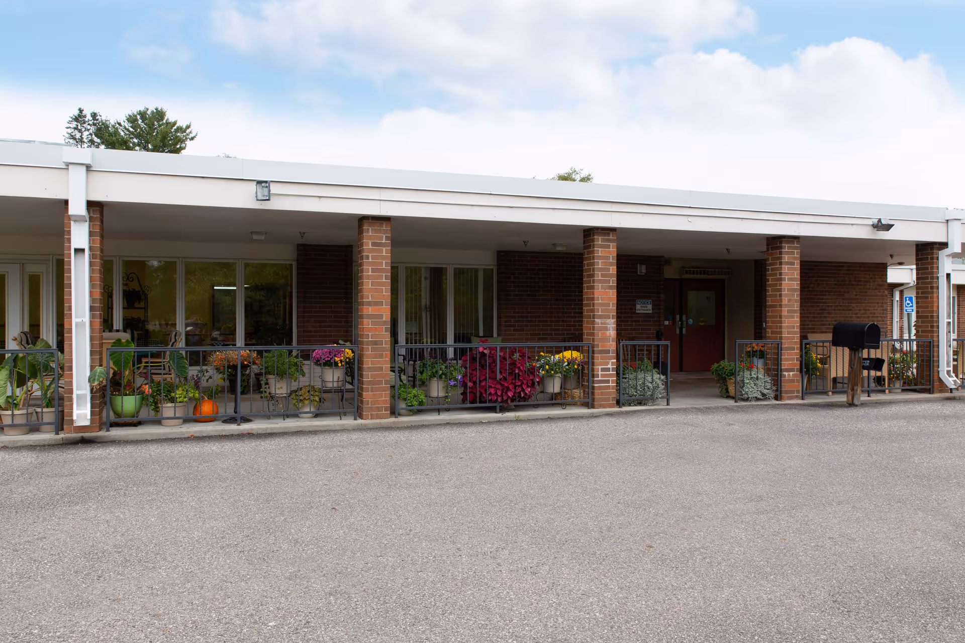 Front entrance of a single-story brick senior living facility with a covered walkway, railings and potted plants.