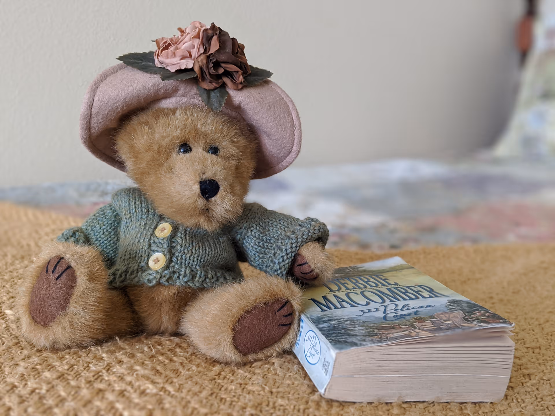 A small teddy bear wearing a knitted sweater and pink hat sits on a bed next to a paperback book.