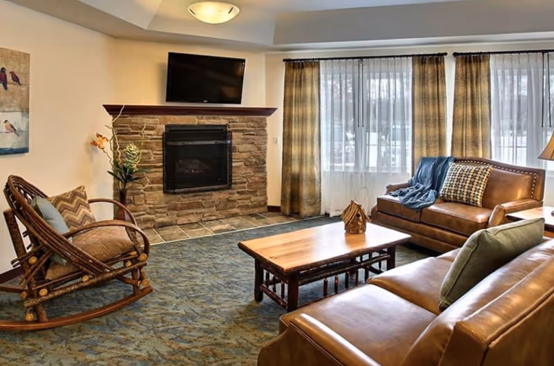 Cozy living room with leather sofas, a rocking chair, wooden coffee table, and a stone fireplace beneath a mounted TV.