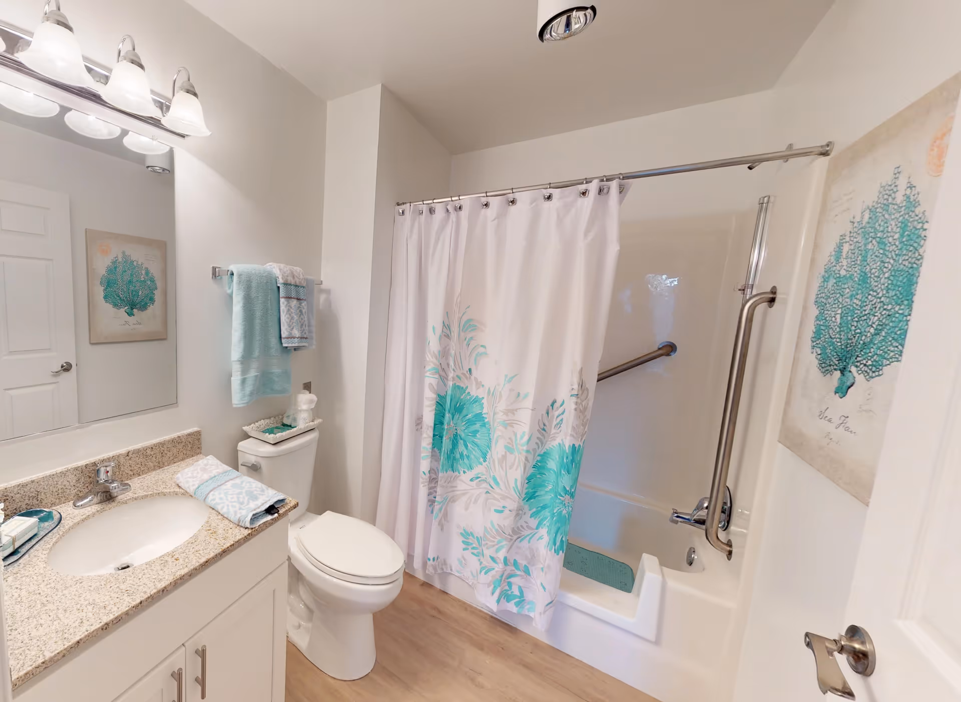 A clean and bright bathroom featuring a white vanity with a granite countertop and an oval sink. Above the sink is a large mirror with a three-light fixture. Next to the vanity is a toilet with a towel rack above it holding folded turquoise and white towels. The bathtub has a white shower curtain with turquoise floral designs and a built-in grab bar for safety. The walls are white, and there is a framed artwork of a turquoise sea fan coral on the wall.