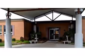 Entrance to a building with a covered driveway supported by columns. The building has a brick exterior and glass doors at the entrance. There are plants and small bushes near the entrance.