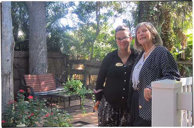 Two women standing together outdoors in a garden area with trees, plants, and a bench visible in the background. One woman is wearing glasses and a black top, and the other woman is wearing a polka dot jacket and a white scarf.