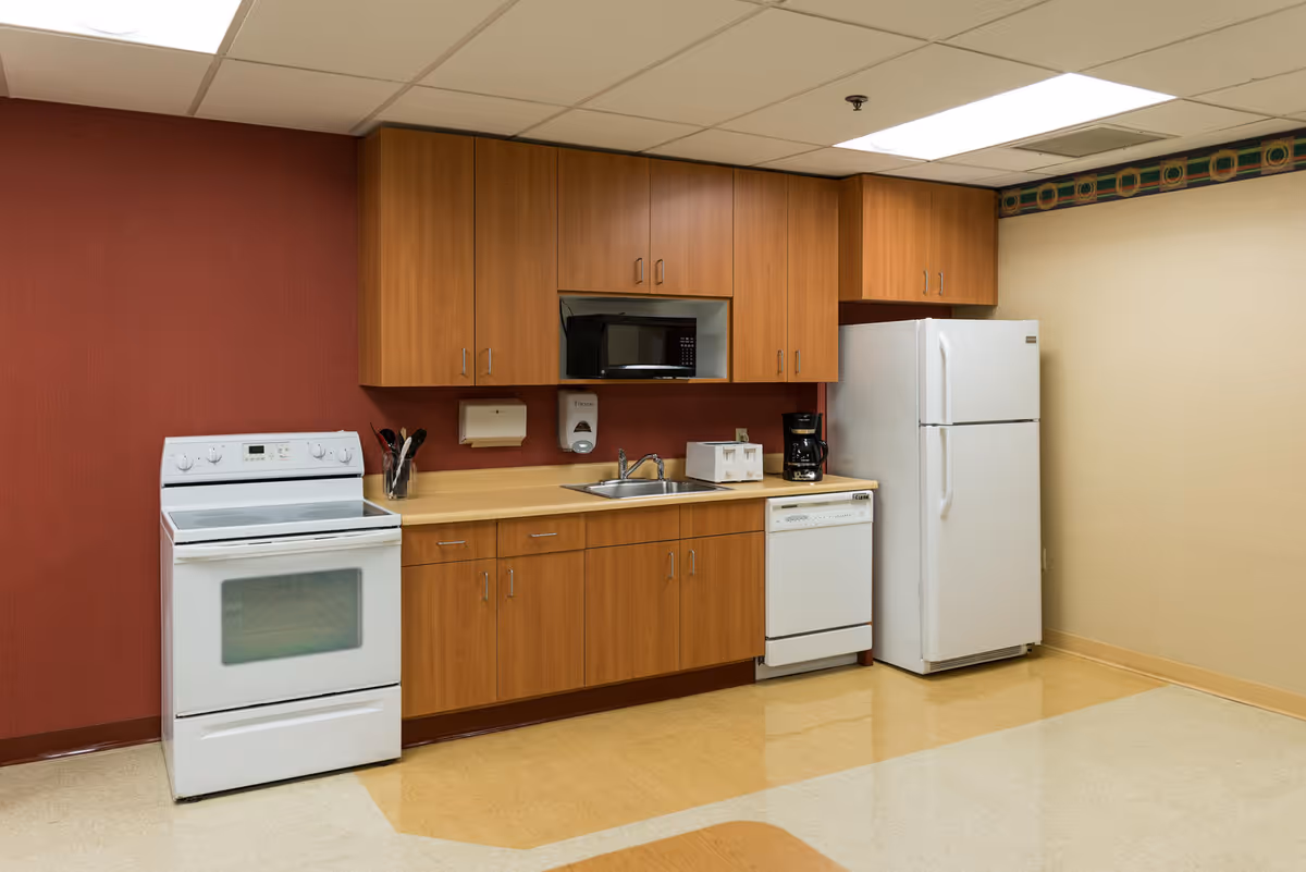 Small institutional kitchenette with a stove, microwave, sink, dishwasher, refrigerator and wood cabinets against a red and beige wall.