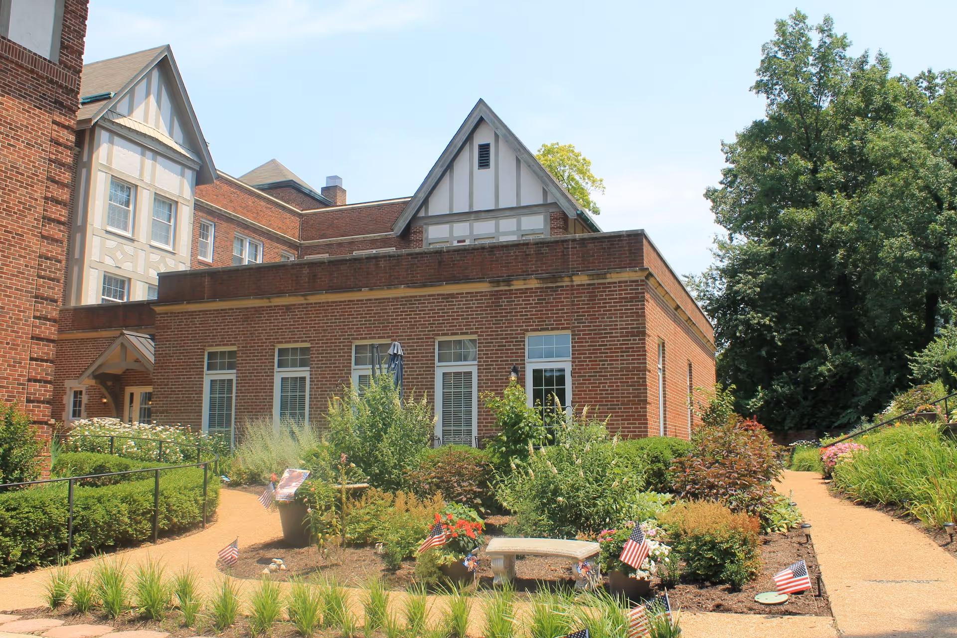 Exterior view of a brick building with white-trimmed windows and a garden area in front. The garden has various plants, flowers, small American flags, and a stone bench. There are paved walkways on either side of the garden, and large trees are visible in the background under a clear sky.