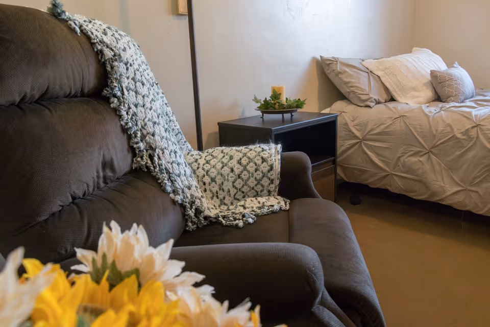 A cozy room with a dark brown armchair draped with a patterned throw blanket, a small dark wooden nightstand with a decorative candle and greenery, and a bed with beige bedding and multiple pillows against a light-colored wall.