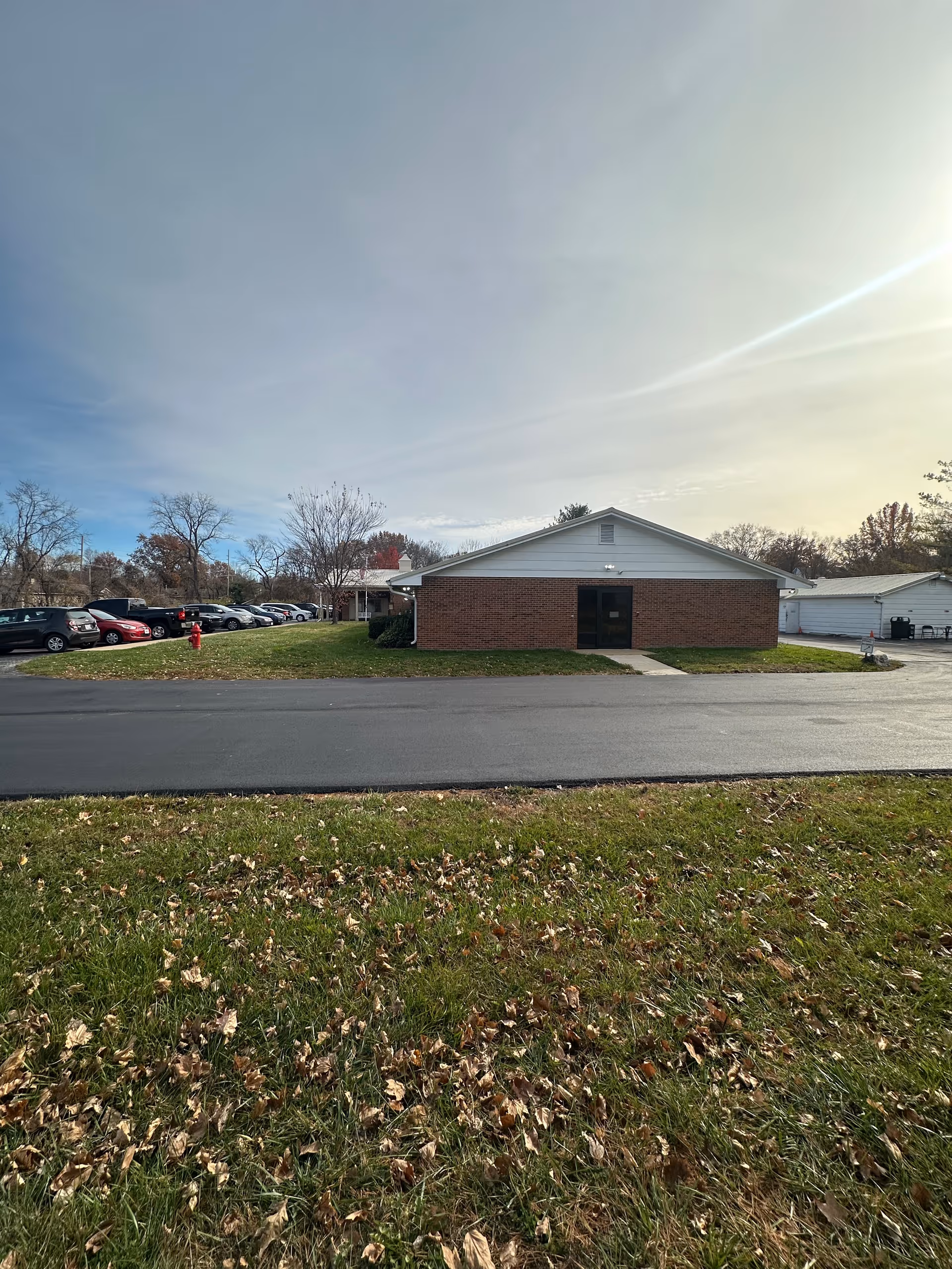 Exterior view of a single-story brick building with a white gabled roof, a paved driveway in front, and a grassy area with fallen leaves in the foreground. Several parked cars and leafless trees are visible in the background under a partly cloudy sky.