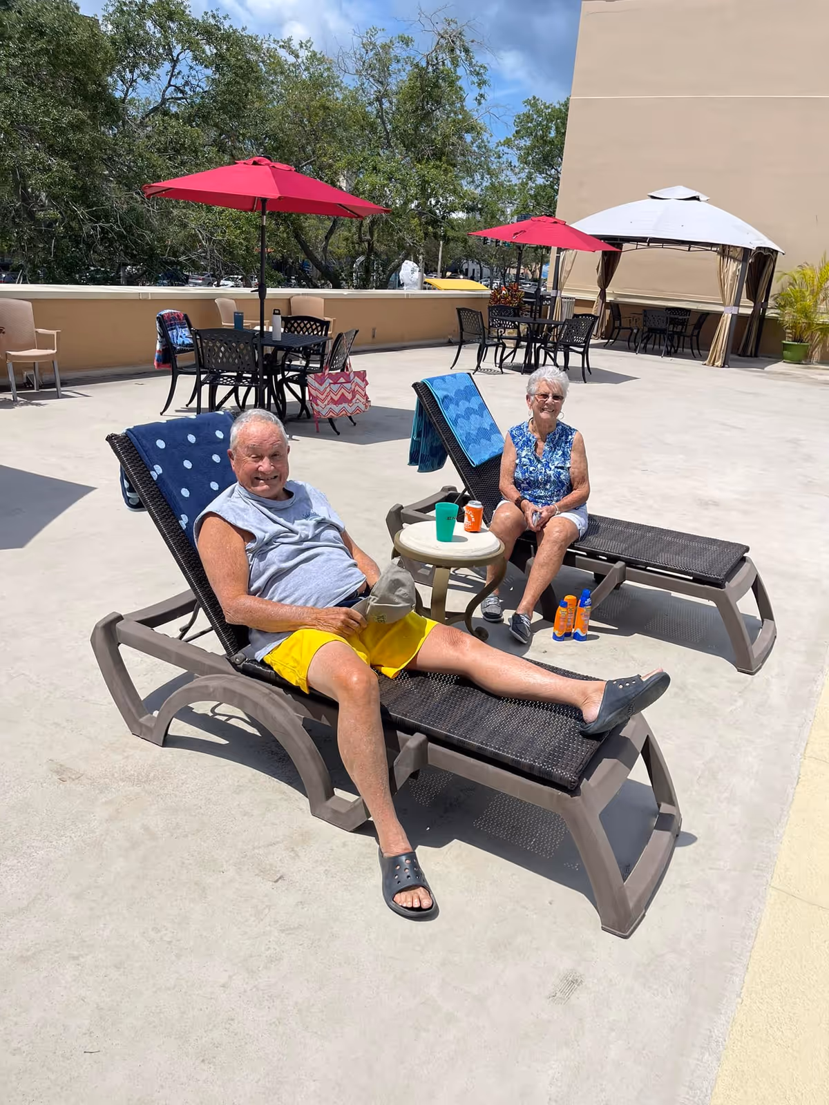 Two elderly people relaxing on lounge chairs on a sunny outdoor patio. One man is wearing a gray sleeveless shirt and yellow shorts, and a woman is wearing a blue patterned sleeveless top and white shorts. There are red umbrellas, tables, and chairs in the background along with some trees and a beige building wall.