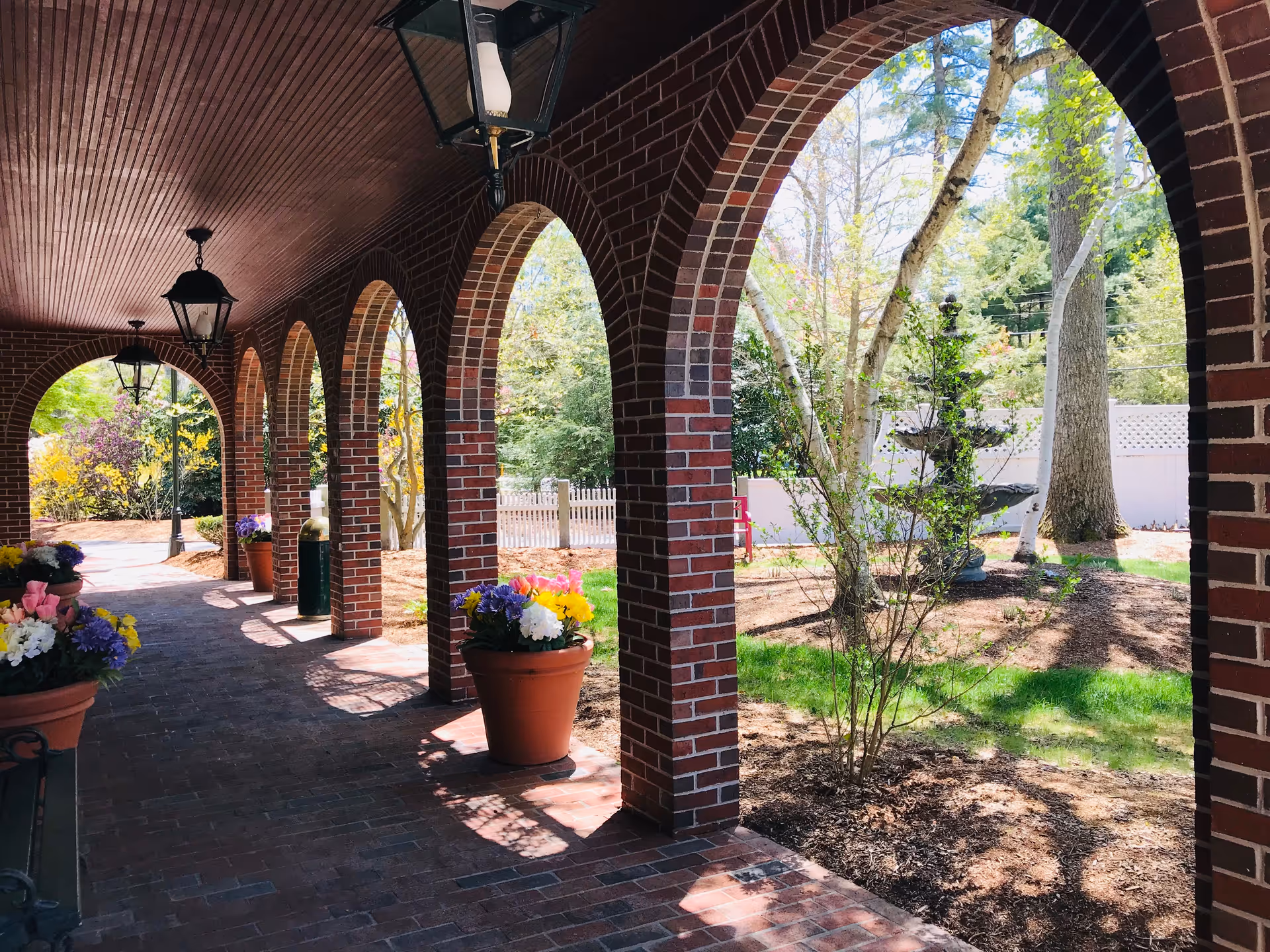 A brick archway corridor with hanging lanterns and large flower pots filled with colorful flowers. The corridor overlooks a garden area with trees, a white fence, and a multi-tiered stone fountain.