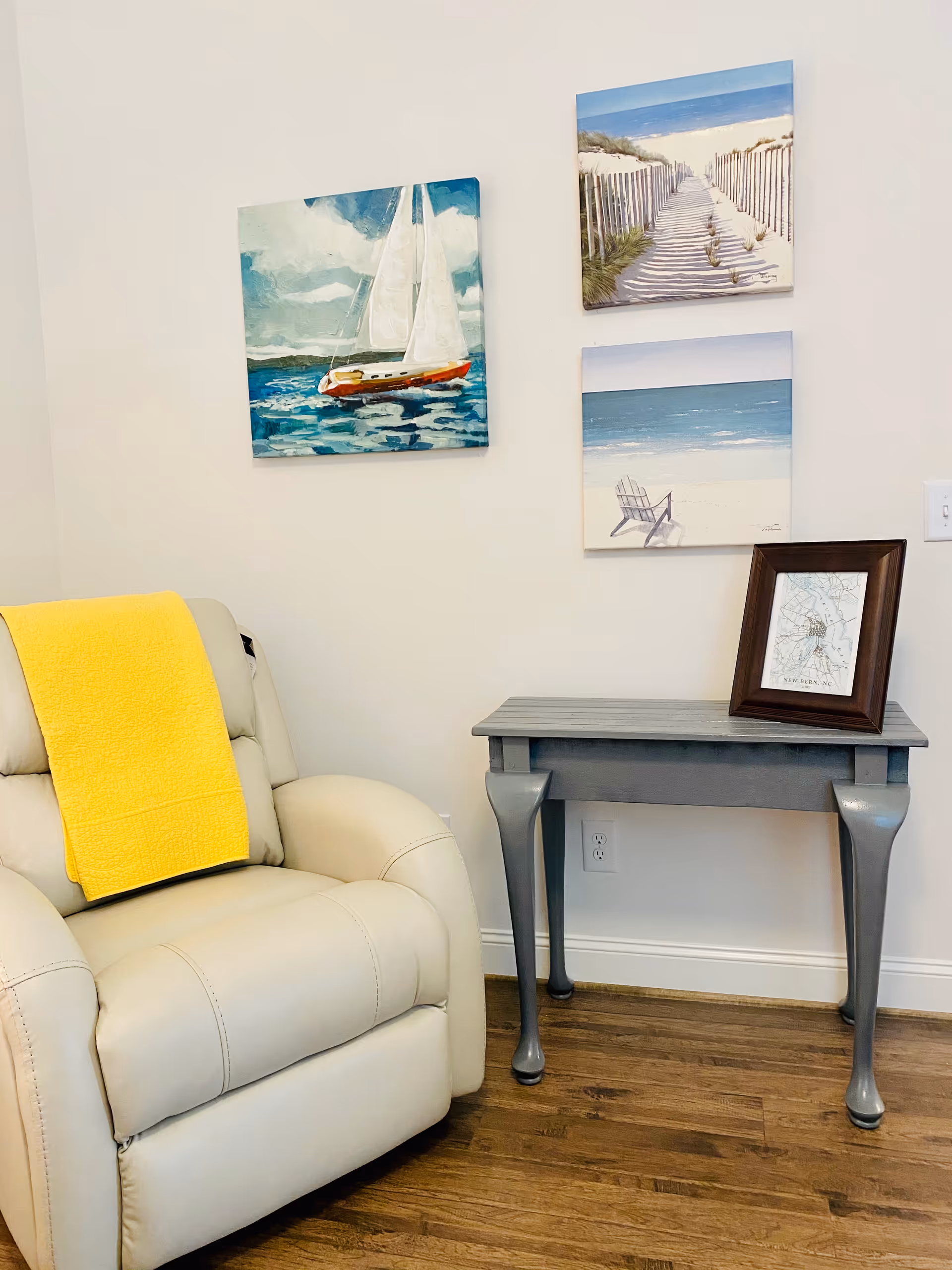 A cozy corner of a room featuring a beige recliner chair with a bright yellow blanket draped over the back. Next to the chair is a small gray wooden table with curved legs, holding a framed map. On the wall above the table are three paintings depicting nautical and beach scenes: a sailboat on the water, a sandy path with a fence leading to the beach, and a beach chair facing the ocean. The room has wooden flooring and white walls.