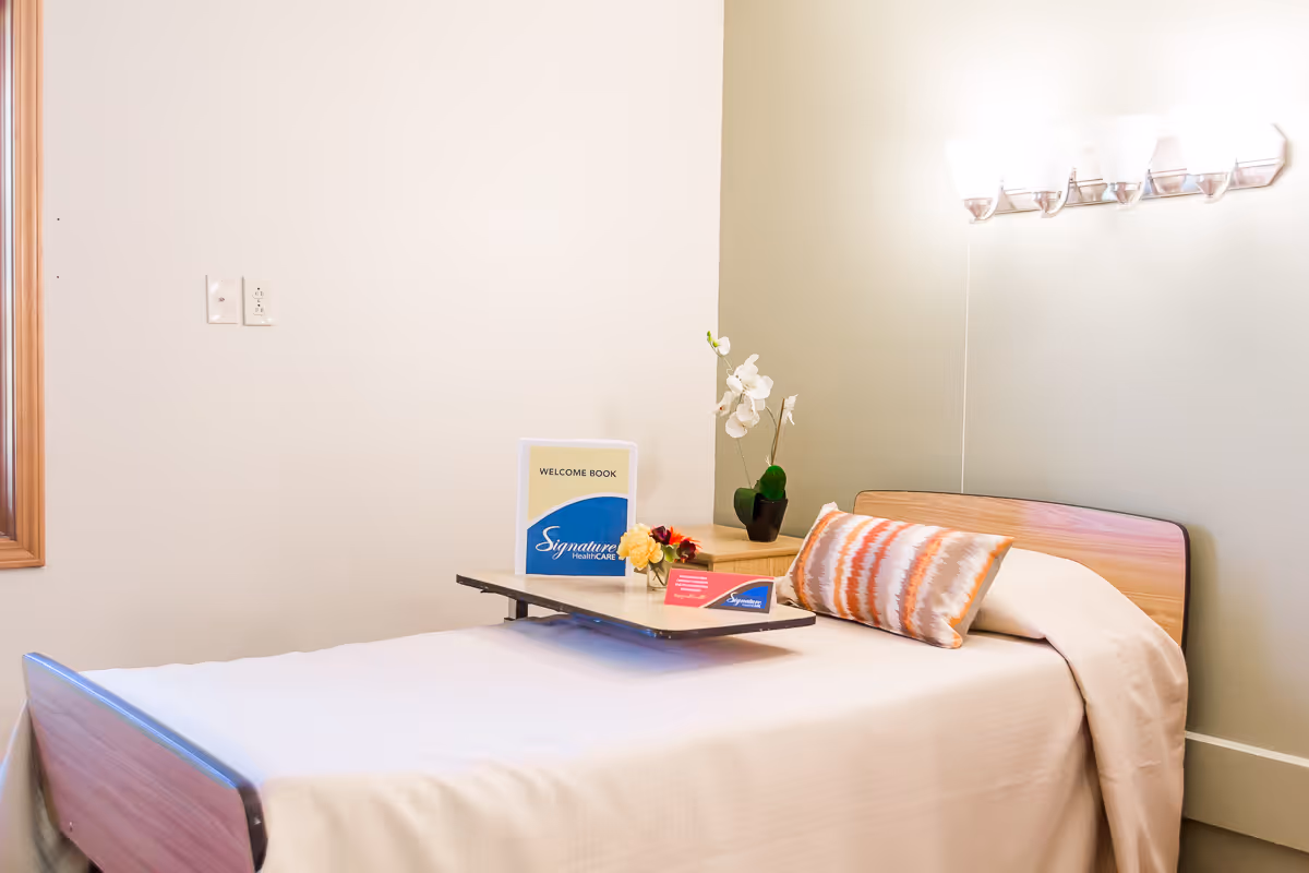 A neatly made single bed in a rehab room with a bedside table holding a welcome book, flowers and a decorative pillow under a wall light.