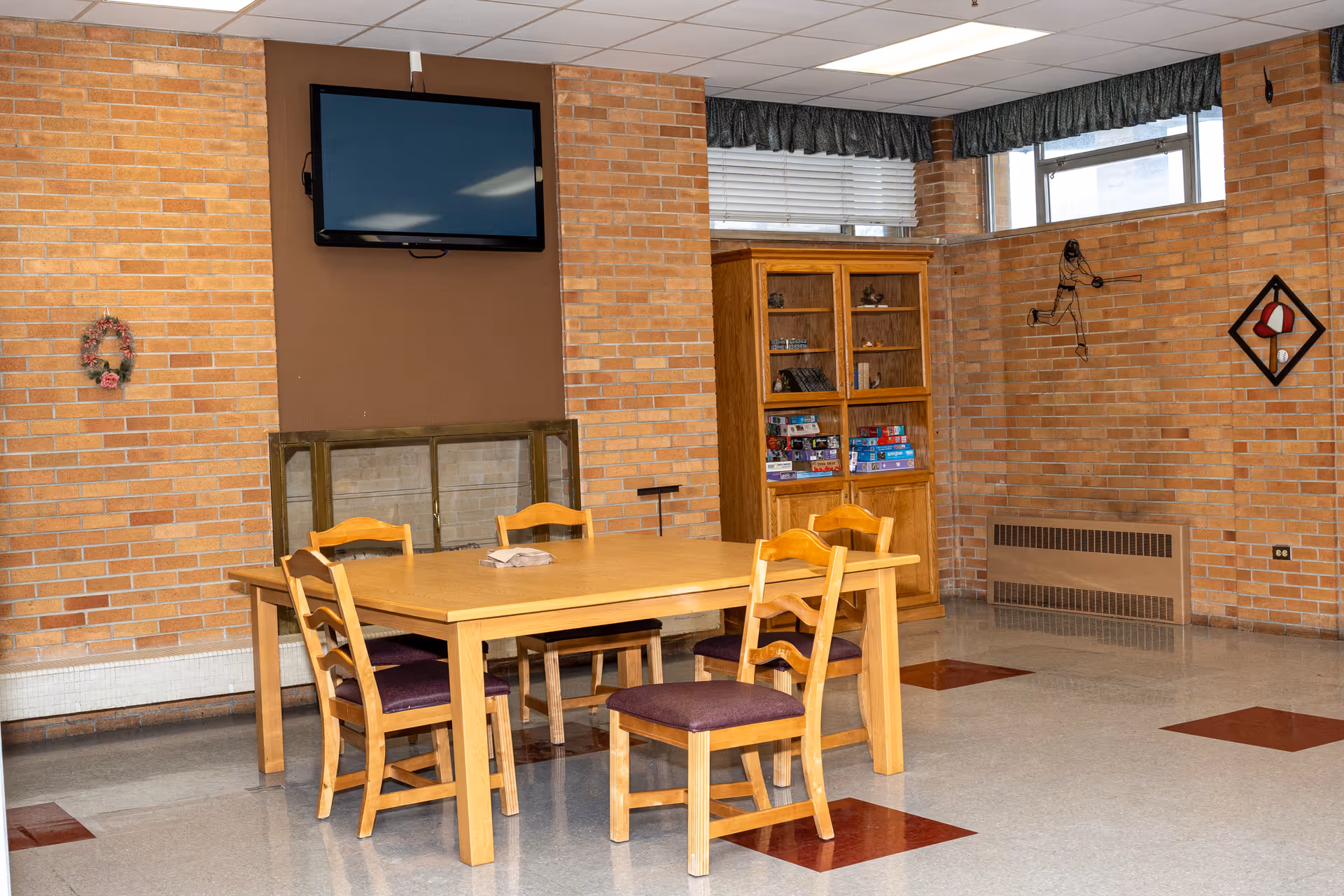 A common area with a wooden table and five chairs with purple cushions. The room has brick walls, a mounted flat-screen TV above a fireplace, a wooden cabinet filled with board games, and wall decorations including a wreath and a baseball-themed metal art piece. The floor is tiled with a pattern of red and gray squares.