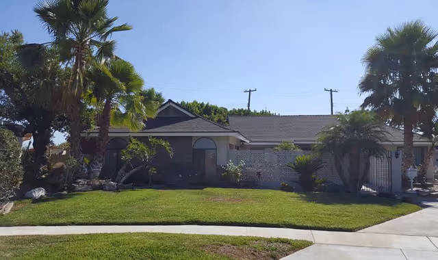 Single-story building with a dark roof and light-colored walls surrounded by palm trees and green grass under a clear blue sky.