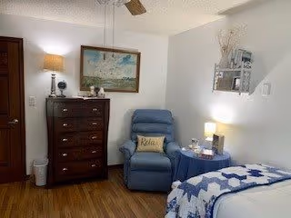 A cozy bedroom featuring a wooden chest of drawers with decorative items on top, a blue recliner chair with a 'Relax' pillow, a small round table with a lamp and other items, and a bed with a blue and white quilt. The walls are light-colored, and there is a ceiling fan above.