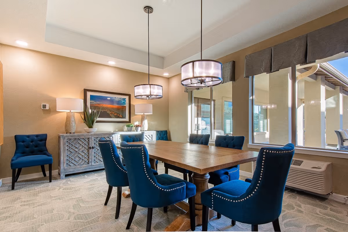 Light-filled dining room with a wooden table surrounded by blue upholstered chairs, pendant lights, a sideboard and large windows.