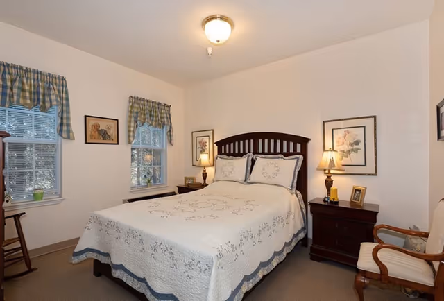 A cozy bedroom in an assisted living facility featuring a double bed with a white and blue floral quilt, two bedside tables with lamps, framed floral artwork on the walls, two windows with blue and green plaid valances, and a wooden armchair with a cushioned seat.