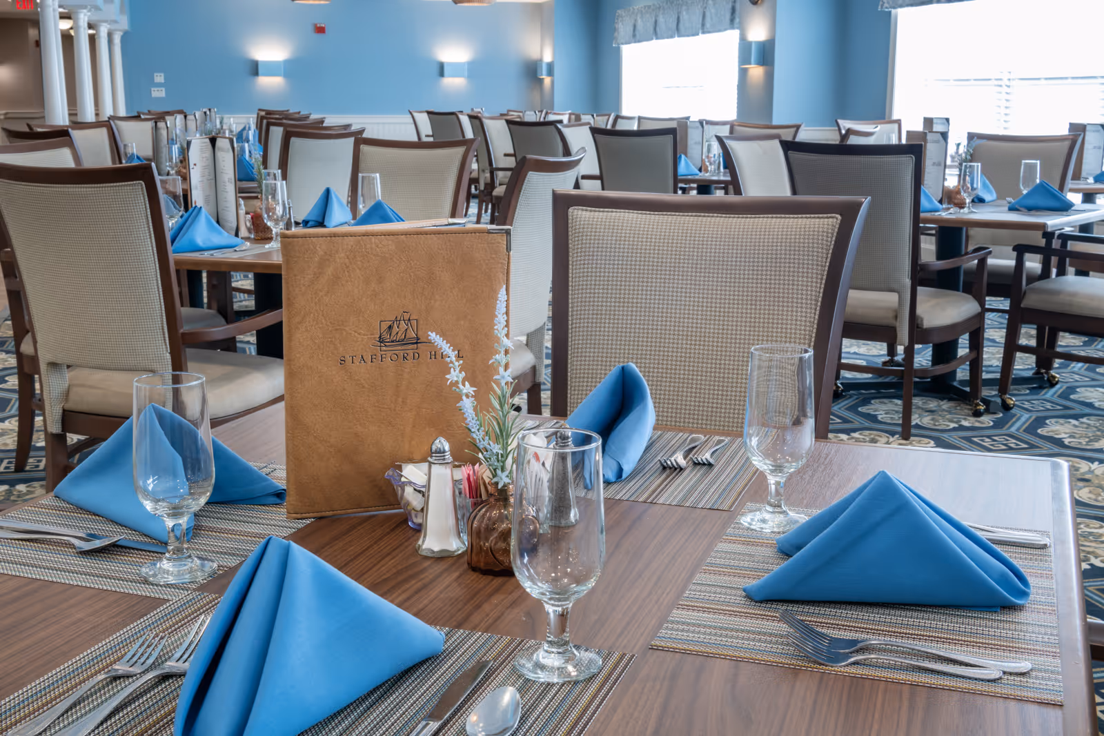 A dining room with multiple tables set with blue folded napkins, clear drinking glasses, silverware, and placemats. One table in the foreground has a brown menu with the Stafford Hill logo, a small vase with flowers, and salt and pepper shakers. The room has light blue walls and cushioned chairs with wooden frames.