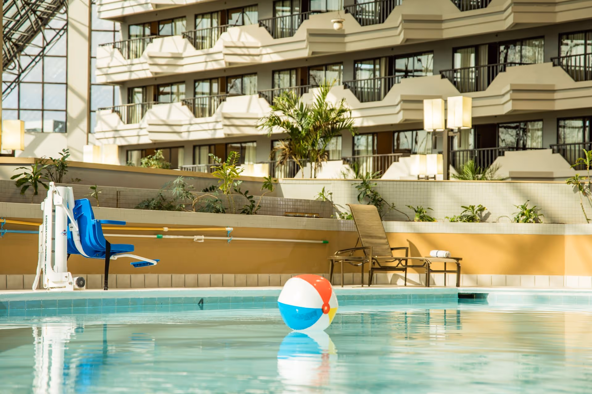 Indoor swimming pool with a colorful beach ball floating on the water, a blue pool lift chair on the left side, and a lounge chair with a rolled towel on the right side. The background shows multiple balconies and large windows of the building interior with plants and soft lighting.