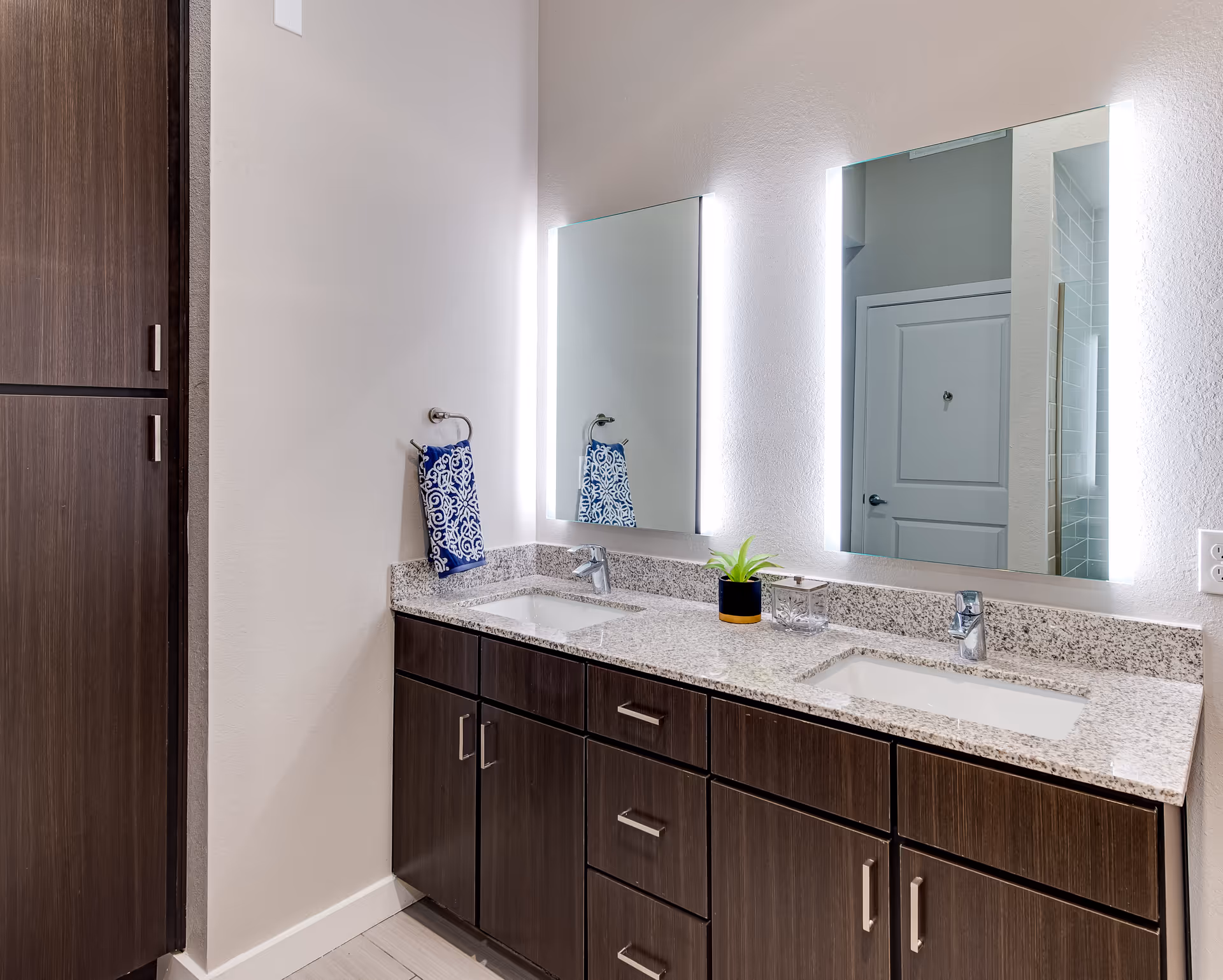 Modern bathroom with a double sink vanity featuring granite countertops and dark wood cabinets. Two rectangular mirrors with built-in lighting are mounted above each sink. A blue and white patterned hand towel hangs on a ring on the wall, and a small green plant and a glass container are placed on the countertop. A closed white door and part of a tiled shower are visible in the reflection of the mirrors.