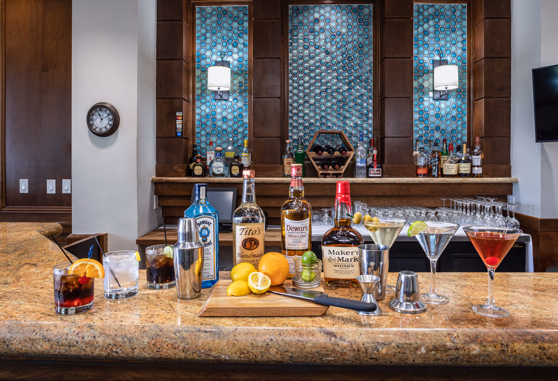 Bar countertop with liquor bottles, cocktail glasses, garnishes and a cutting board set against a decorative tiled back bar.