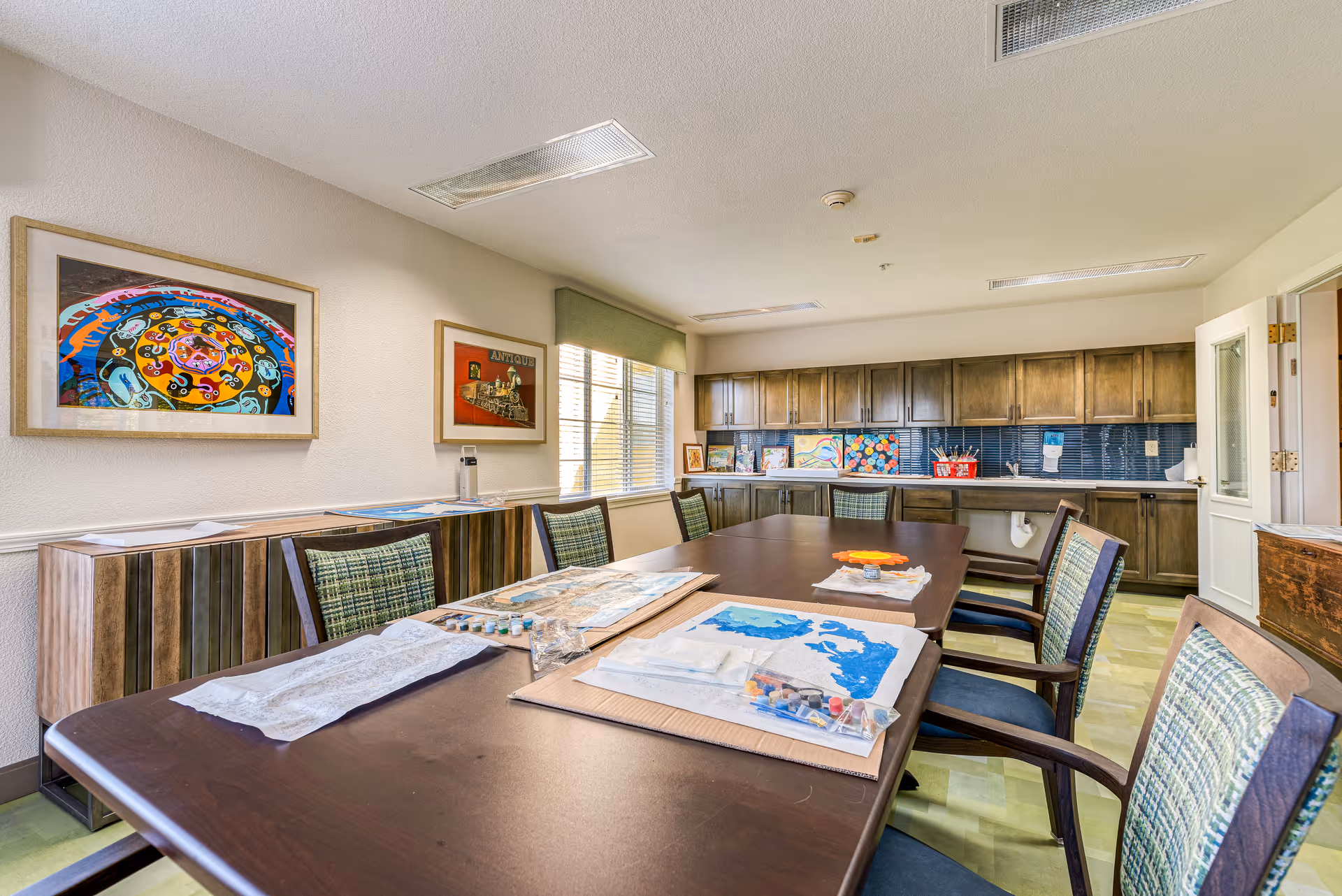 A bright and spacious activity room with a large wooden table surrounded by chairs. On the table are art supplies and partially completed paintings. The room has wooden cabinets along the back wall, colorful artwork displayed on the walls and countertops, and a window letting in natural light.