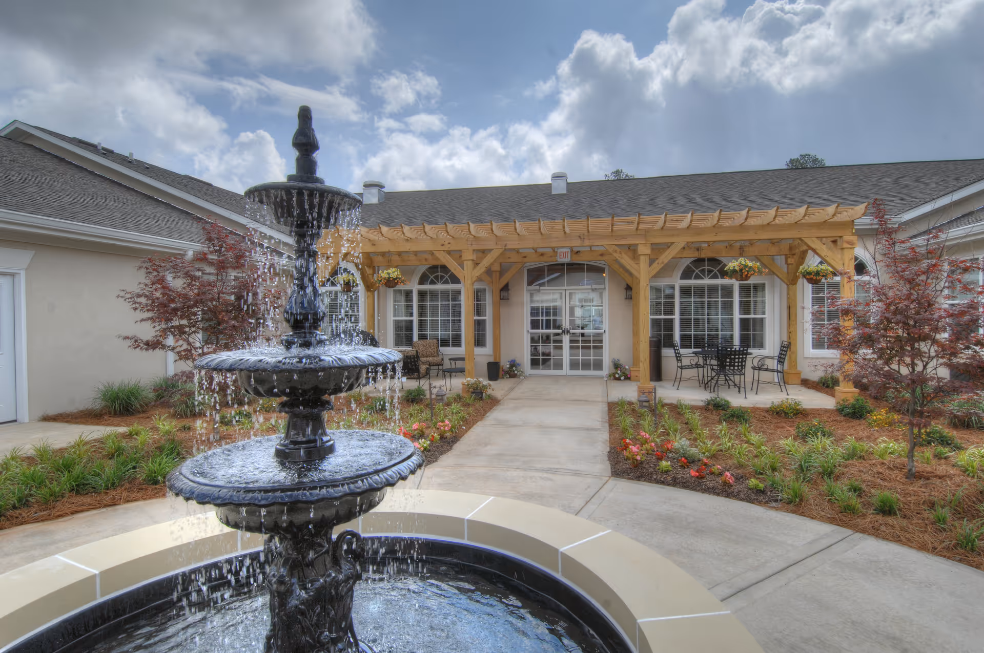 A tiered fountain in a landscaped courtyard leading to a pergola-covered front entrance of a senior living building.