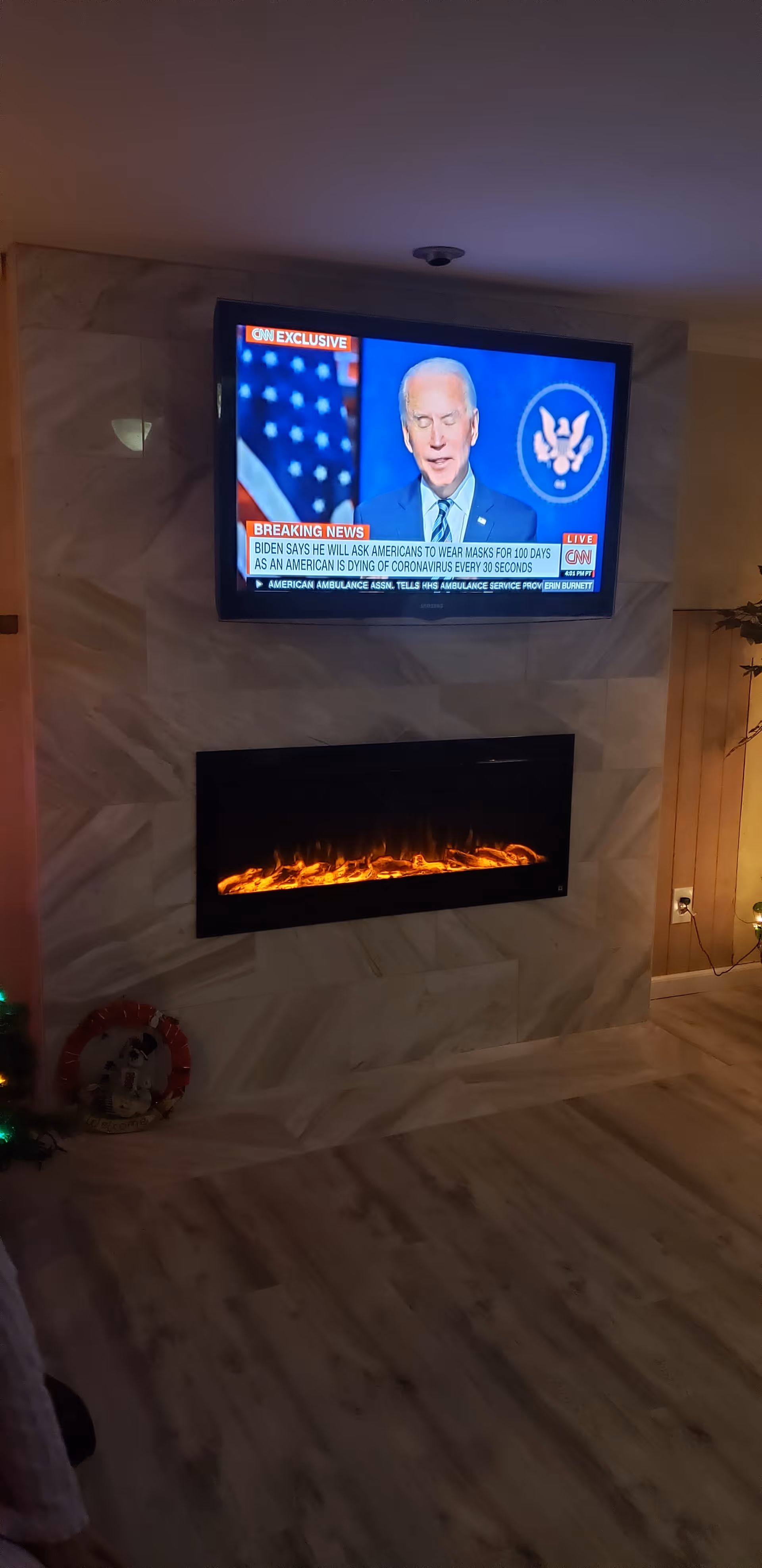 A wall-mounted flat screen TV displaying a CNN news broadcast with Joe Biden speaking. Below the TV is a modern electric fireplace with glowing flames. The wall has a marble-like finish and the floor is light wood. There is a small Christmas decoration with a wreath and a figurine on the floor to the left.