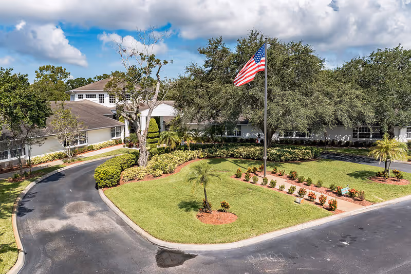View of a senior living facility named The Landmark showing a curved driveway around a landscaped green area with an American flag on a flagpole, surrounded by trees and bushes under a partly cloudy sky.