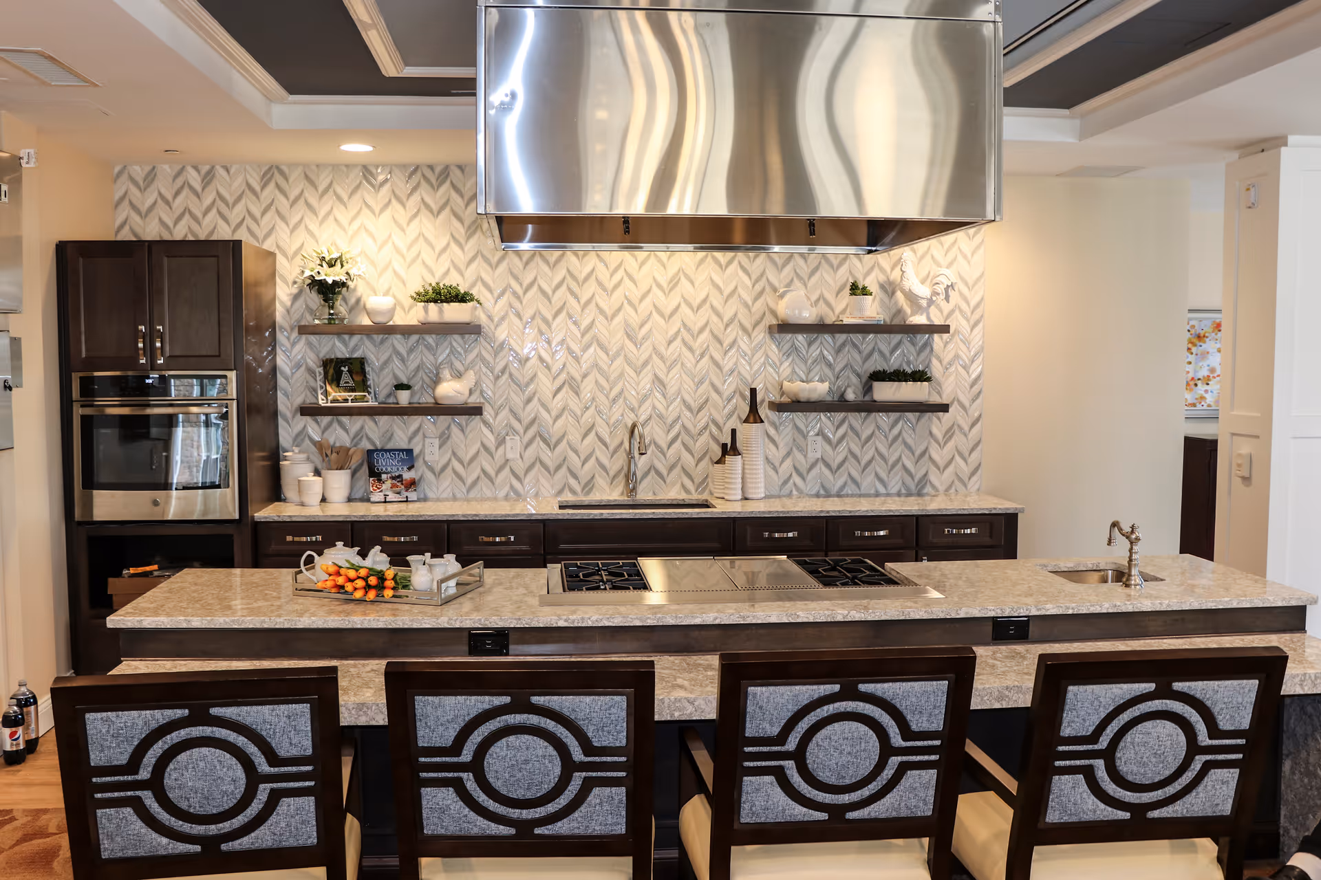 Modern kitchen with a large island countertop featuring a built-in stove and a small sink. Four chairs with decorative backs are positioned at the island. The kitchen has dark wood cabinets, stainless steel appliances, and a patterned backsplash with floating shelves holding decorative items and plants. A large stainless steel range hood is mounted above the stove.