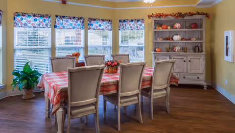 Dining room featuring a long table with a red plaid tablecloth and eight upholstered chairs, a decorative hutch with seasonal items, and a row of windows with floral valances.