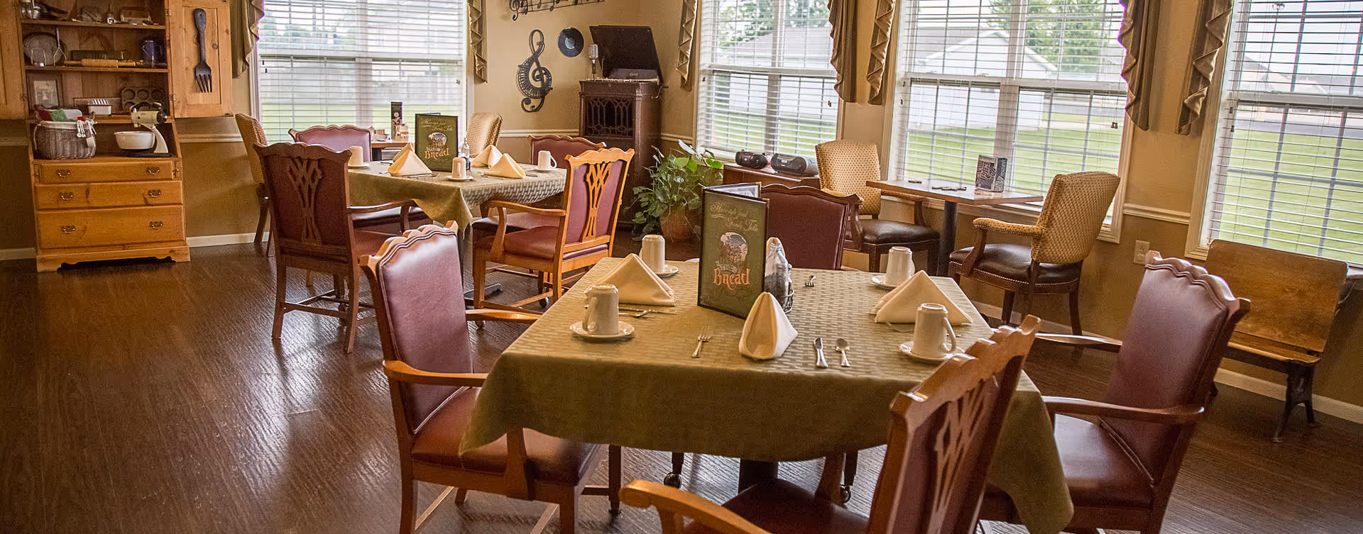 A dining room with wooden floors and several tables covered with green tablecloths. Each table is set with white napkins, cups, and silverware. The room has large windows letting in natural light, wooden chairs with red cushions, and decorative items on the walls and shelves.