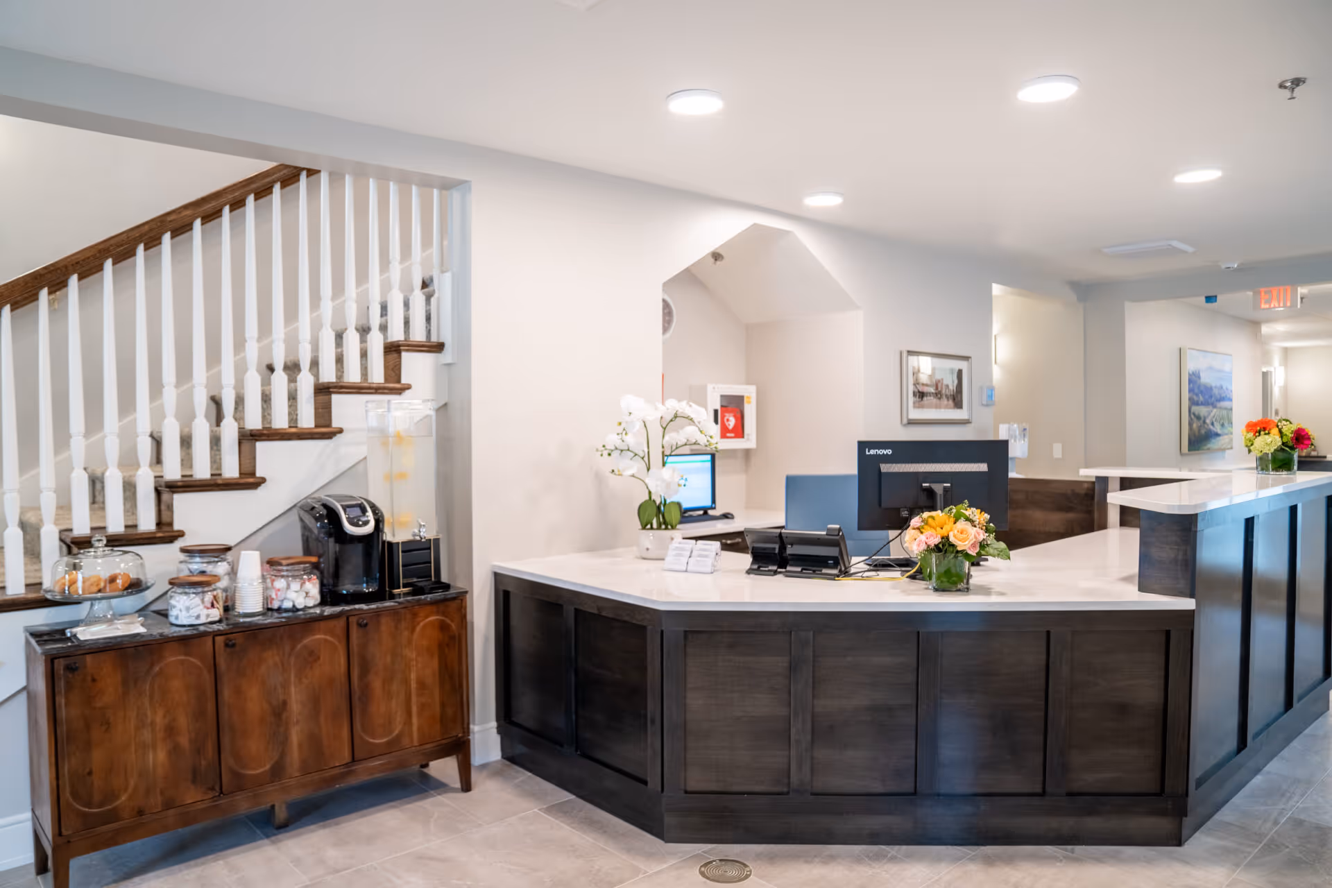 Reception area of Benchmark at Billerica Crossings featuring a dark wood reception desk with a computer monitor and flower arrangements. To the left, there is a wooden sideboard with a coffee maker, water dispenser, and jars of snacks. A staircase with white spindles and wooden handrail is visible behind the sideboard. The space is well-lit with recessed ceiling lights and decorated with framed artwork on the walls.