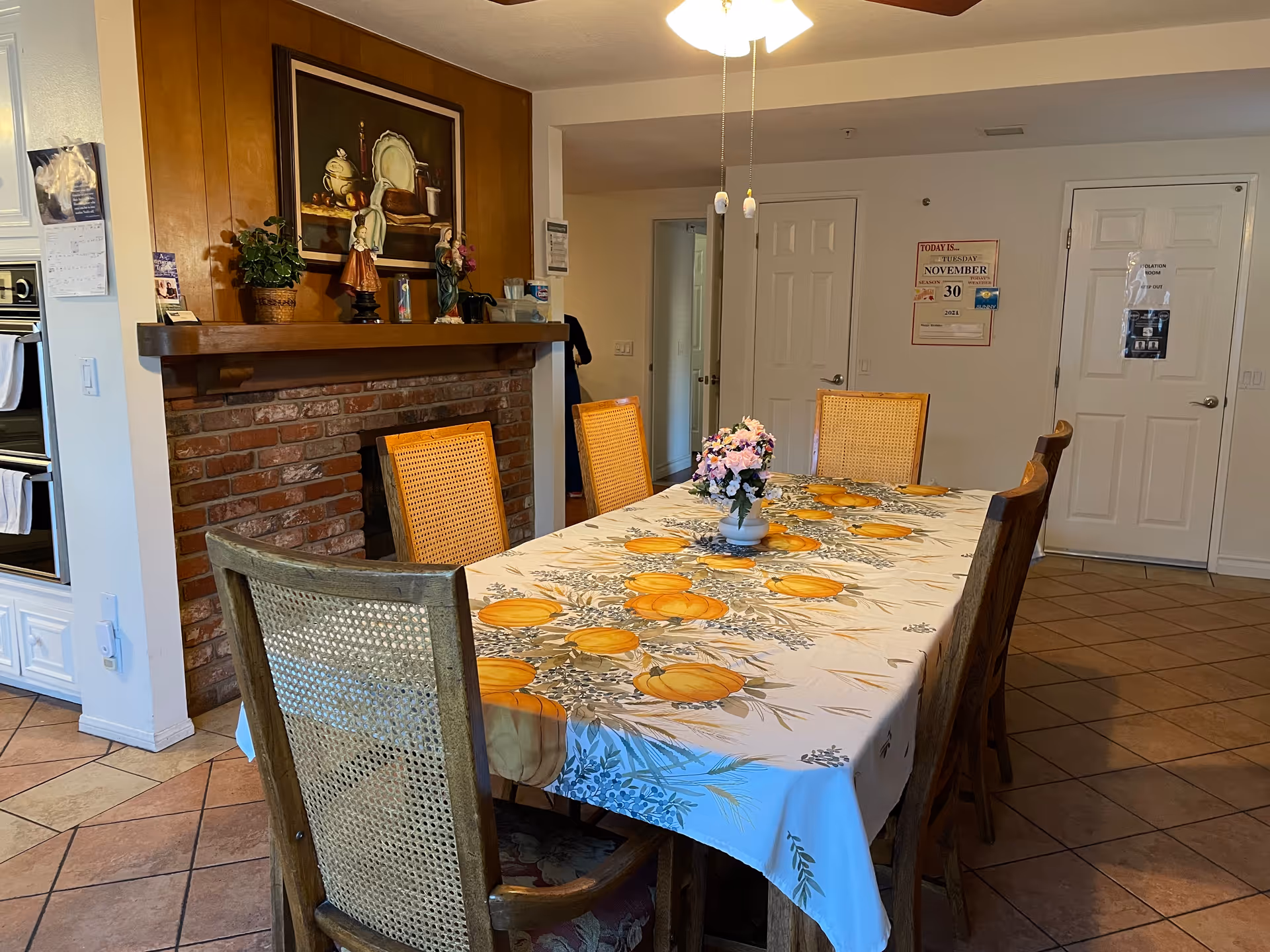 A dining room with a rectangular table covered with a white tablecloth decorated with orange pumpkins and green leaves. Six wooden chairs with woven backs surround the table. A small vase with pink flowers is placed in the center of the table. In the background, there is a brick fireplace with a wooden mantle holding decorative items and a framed painting above it. The room has tiled flooring and white walls with two closed white doors and a ceiling fan with lights above the table.