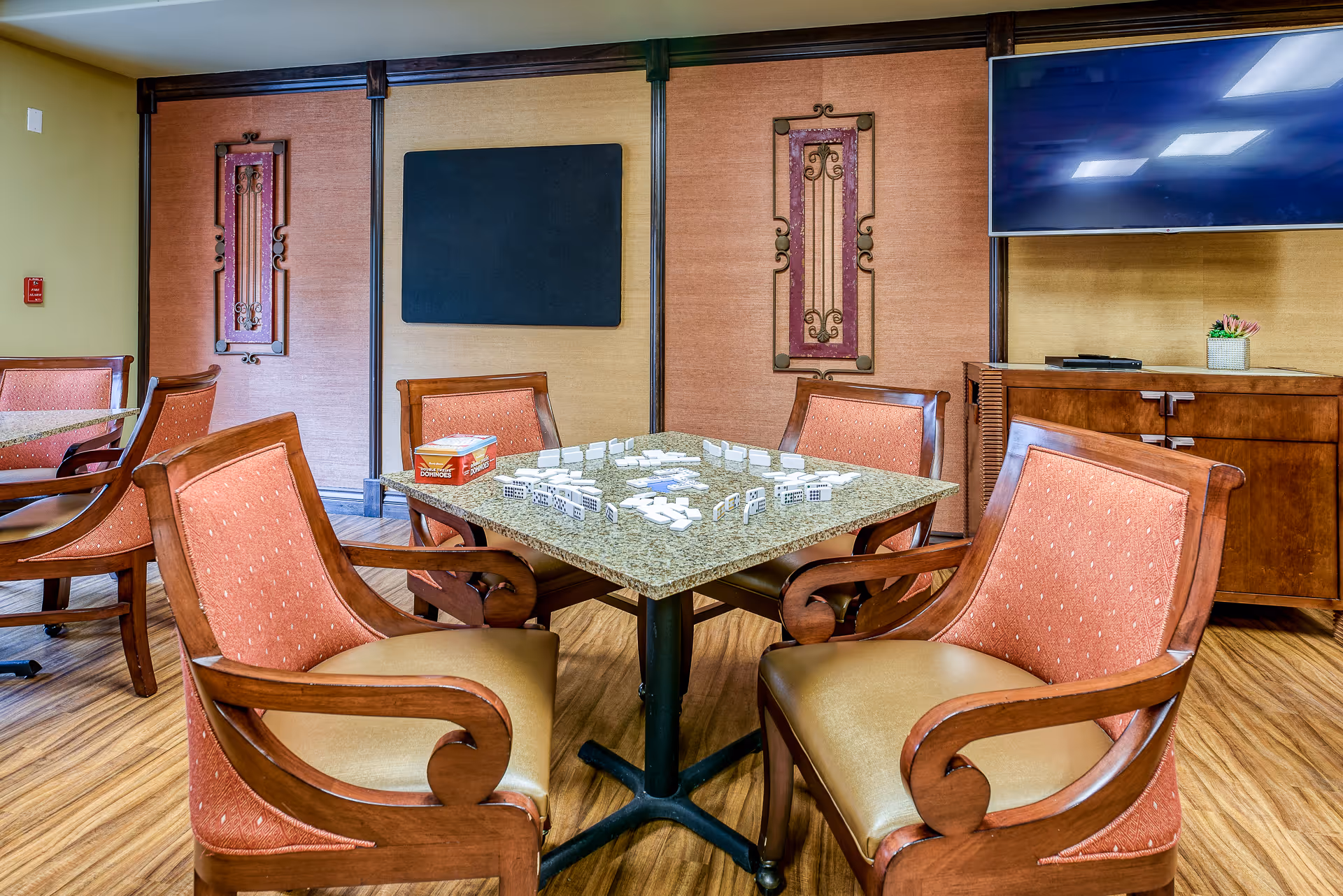 A communal activity room with a square table surrounded by upholstered chairs, game tiles on the table and a large wall-mounted TV.