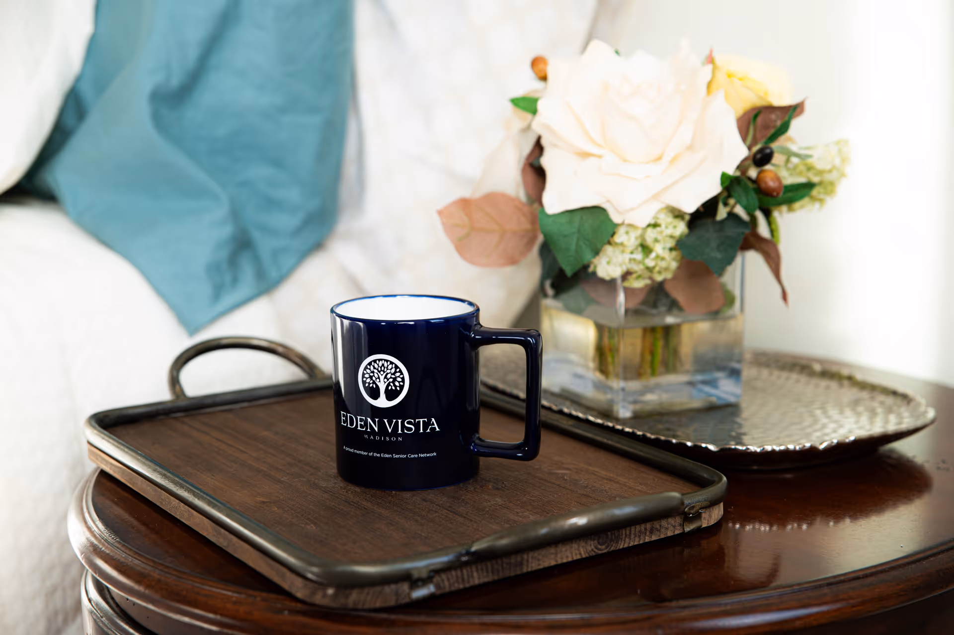 A dark wooden tray on a polished wooden table holding a dark blue mug with the Eden Vista Madison logo. Next to the tray is a silver decorative plate with a square glass vase containing white and yellow flowers with green and brown leaves. A white quilted fabric and a teal cloth are visible in the background.
