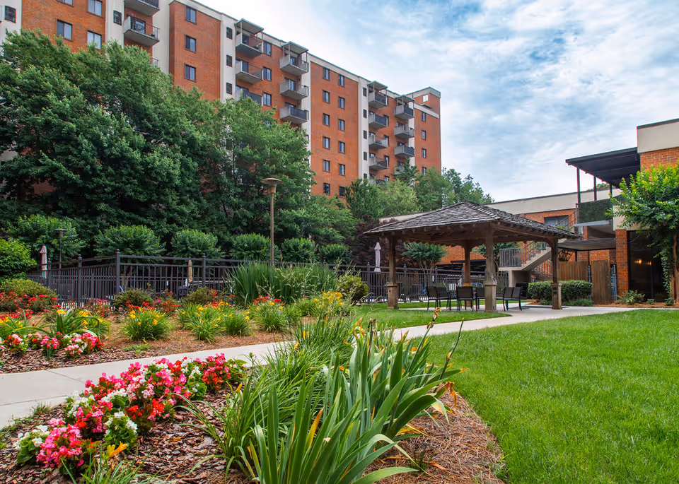 Courtyard with a wooden gazebo, landscaped flower beds and a grassy lawn in front of a multi-story brick apartment building.