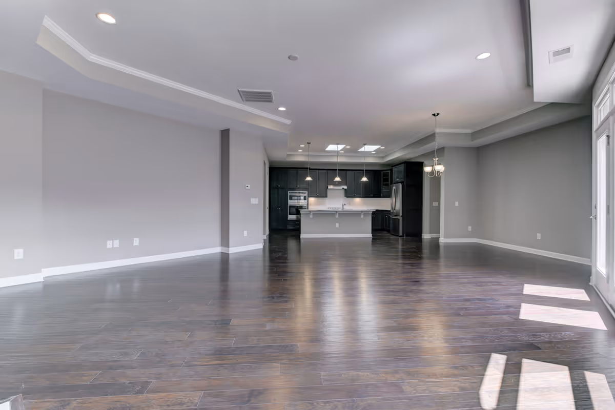 Spacious open floor plan interior with dark hardwood floors, light gray walls, recessed lighting, and a modern kitchen featuring dark cabinetry, stainless steel appliances, and pendant lights over a large island. Large windows on the right side allow natural light to fill the space.