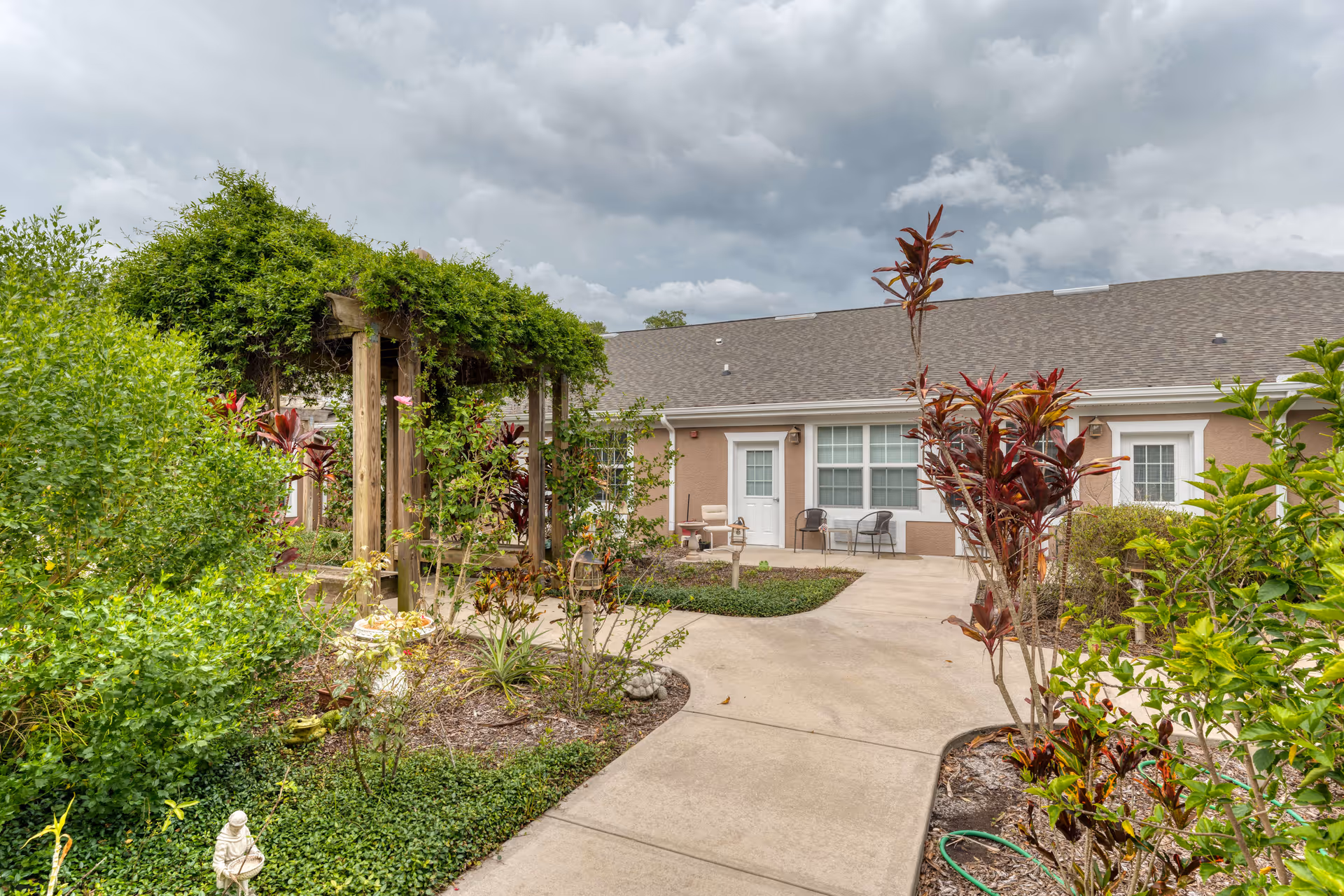 Outdoor garden area at Addington Place of Titusville with a wooden pergola covered in green vines, various plants and shrubs, a concrete pathway, and a building with windows and doors in the background under a cloudy sky.