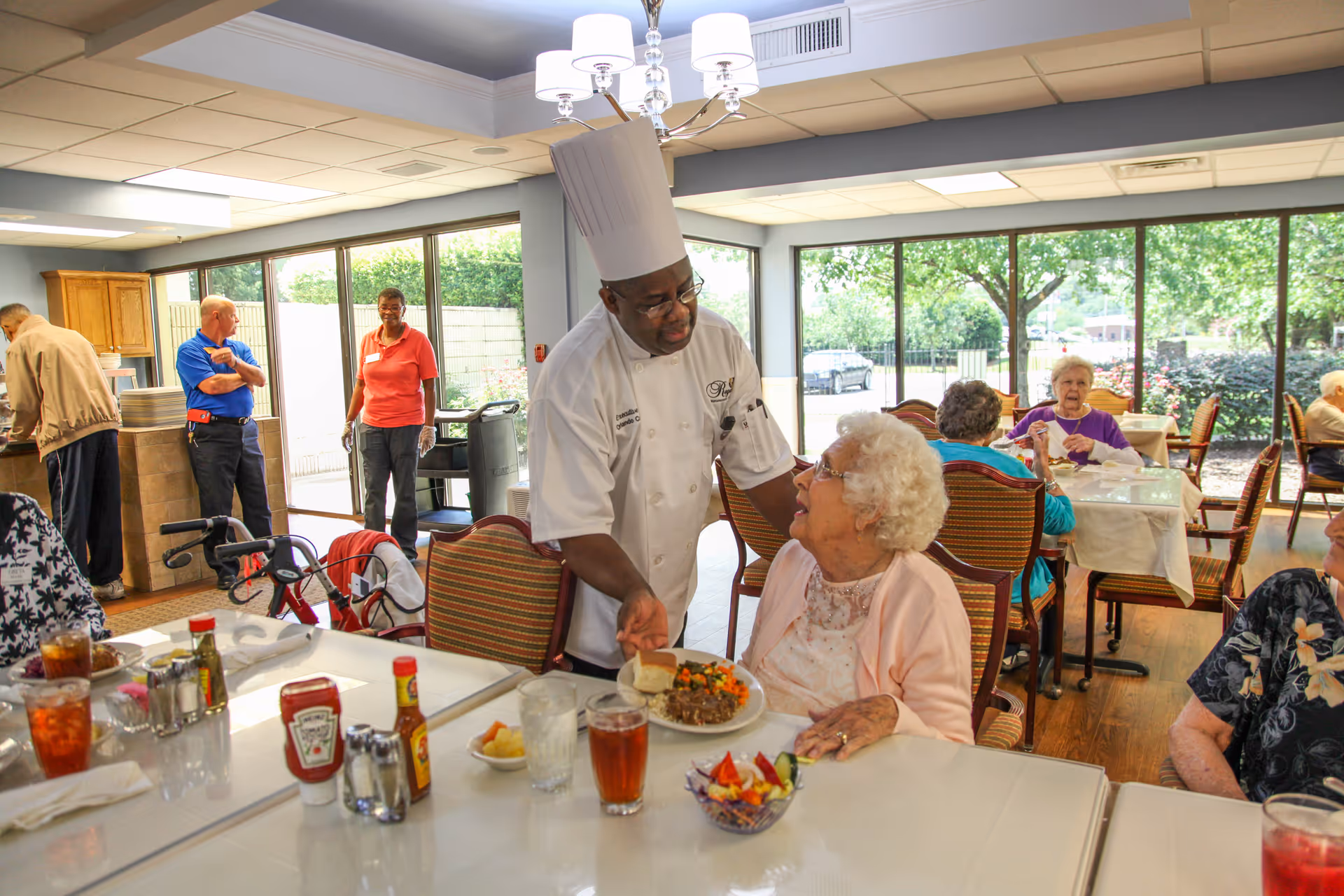 A chef wearing a white uniform and tall chef hat is serving a meal to an elderly woman seated at a dining table in a bright dining room with large windows showing greenery outside. Other elderly residents are seated at tables enjoying their meals, and staff members are visible in the background near the kitchen area.