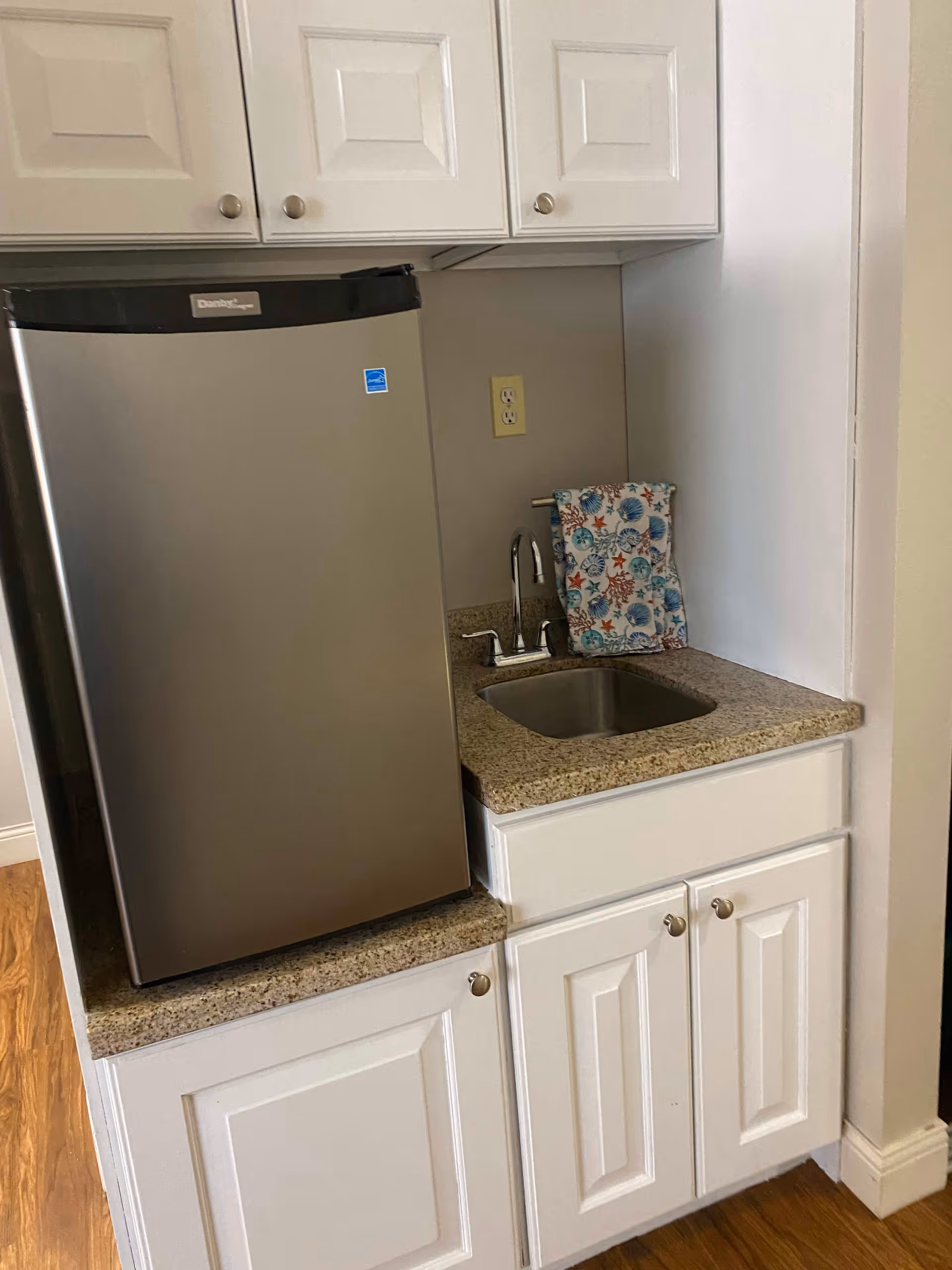 Small kitchen area with a stainless steel mini refrigerator, a granite countertop with a built-in sink, white cabinets above and below the counter, and a colorful towel hanging on a small towel rack next to the sink.