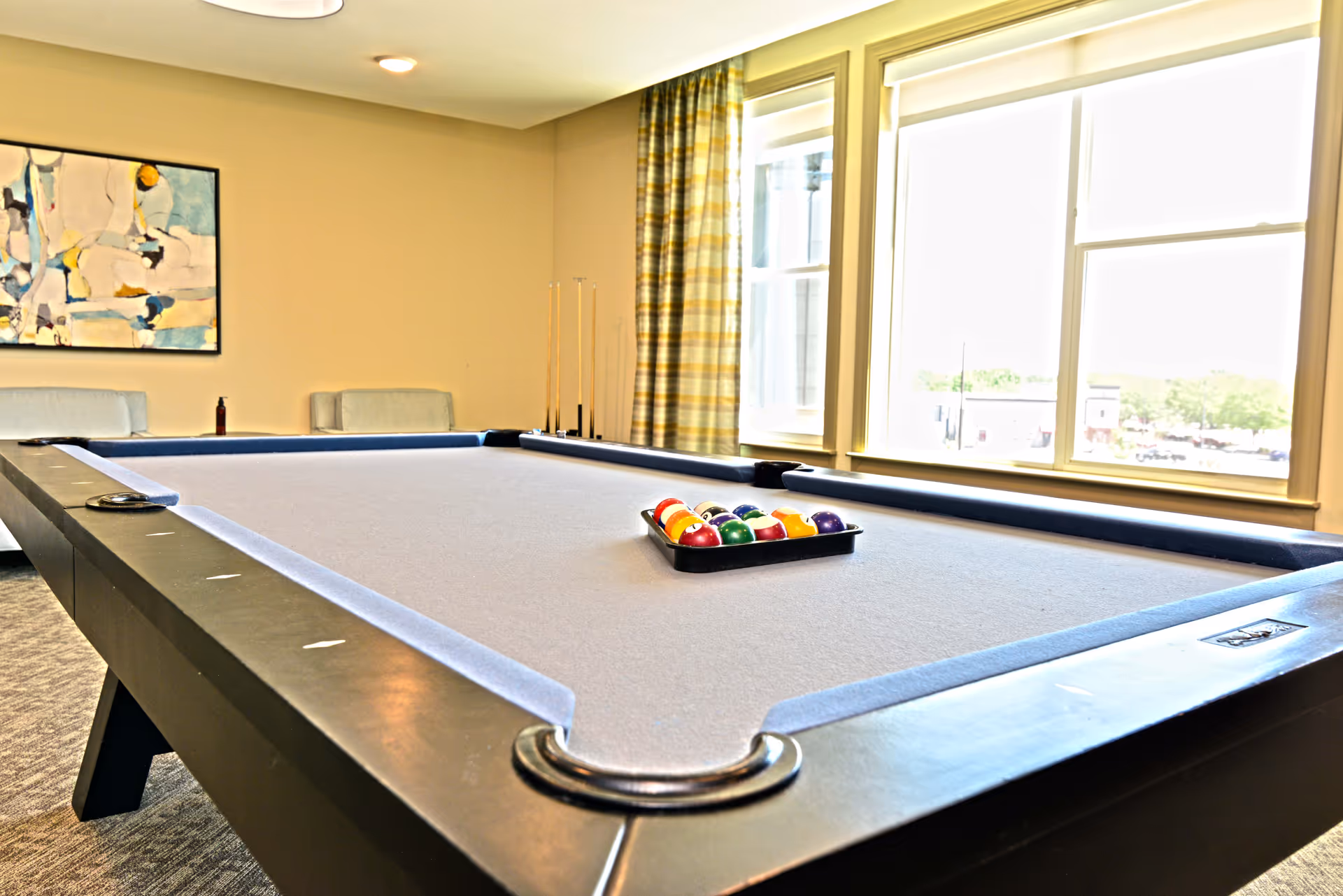 A bright room with a pool table in the center, set up with a rack of billiard balls. The room has large windows with striped curtains letting in natural light, a piece of abstract art on the wall, and seating along the back wall.