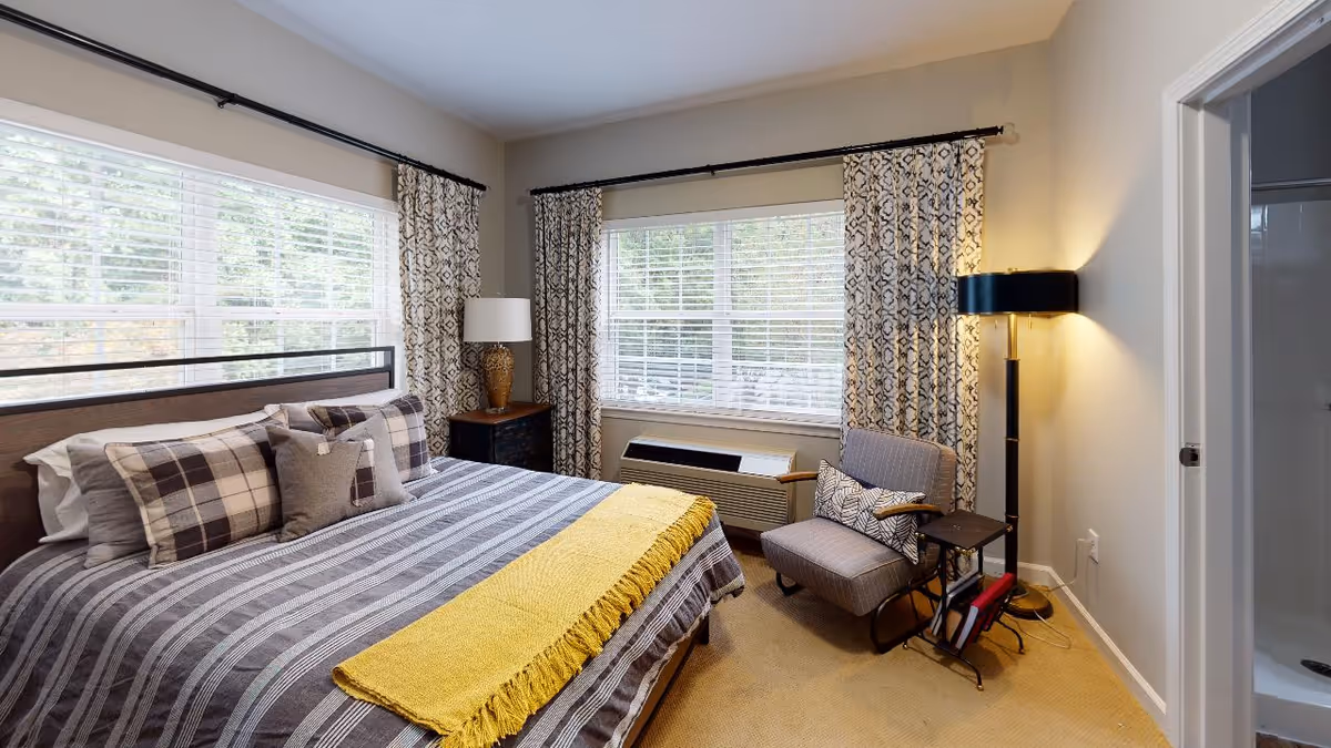 A cozy bedroom with a large bed featuring striped and plaid bedding, a yellow throw blanket, and multiple pillows. The room has two large windows with patterned curtains, a wooden nightstand with a lamp, a cushioned armchair with a patterned pillow, a floor lamp, and a small side table holding books. There is a doorway leading to a bathroom with a visible shower.