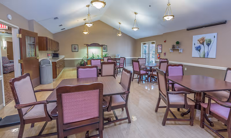 A dining room with several wooden tables and chairs with pink upholstery. The room has a high ceiling with hanging light fixtures, beige walls, and a wooden floor. There is a sideboard with cabinets and a trash can along one wall, a door leading to another room, and a painting of flowers on the wall.