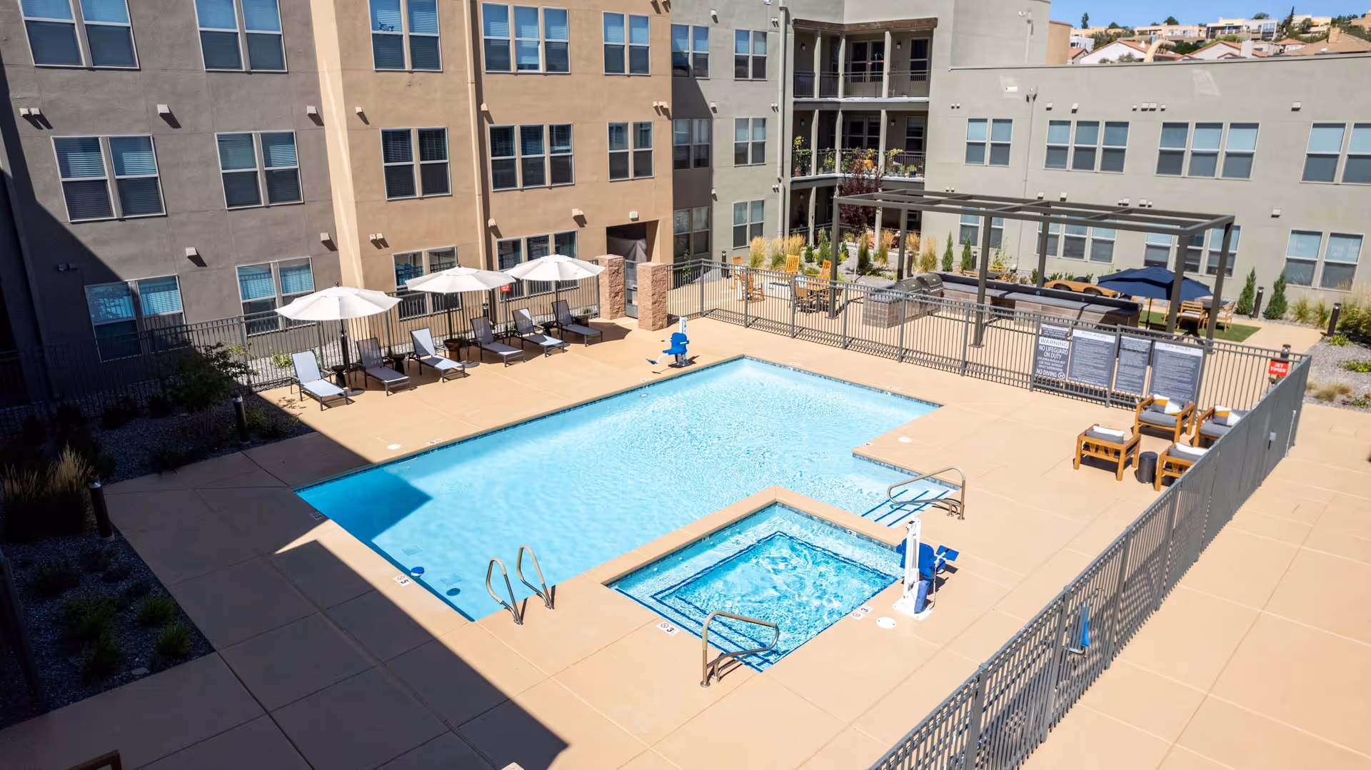 Outdoor swimming pool and hot tub area surrounded by a beige concrete deck with lounge chairs and umbrellas. The pool area is fenced and adjacent to a multi-story residential building with multiple windows. There is a shaded seating area with tables and chairs in the background.