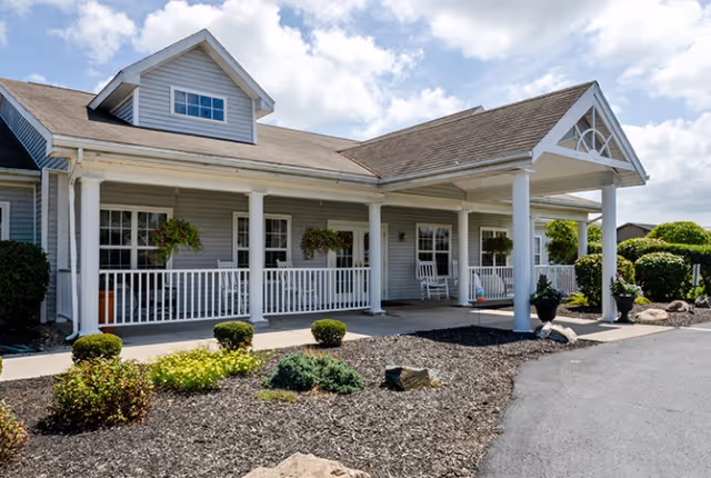 Exterior view of a single-story building with a covered porch supported by white columns. The porch has white railings and hanging flower baskets. There are shrubs and landscaped plants in front of the building under a partly cloudy sky.