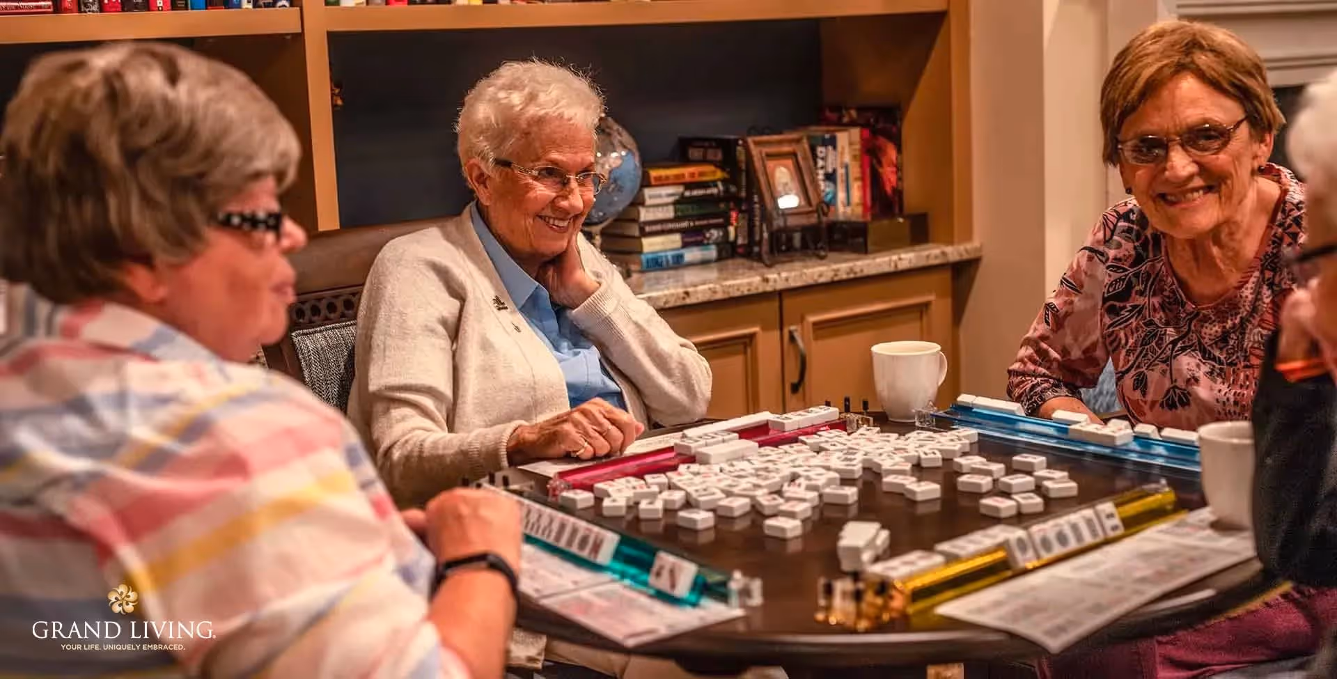 Four elderly women sitting around a table playing a game of Mahjong in a cozy room with bookshelves and cabinets in the background. They are smiling and appear to be enjoying their time together.