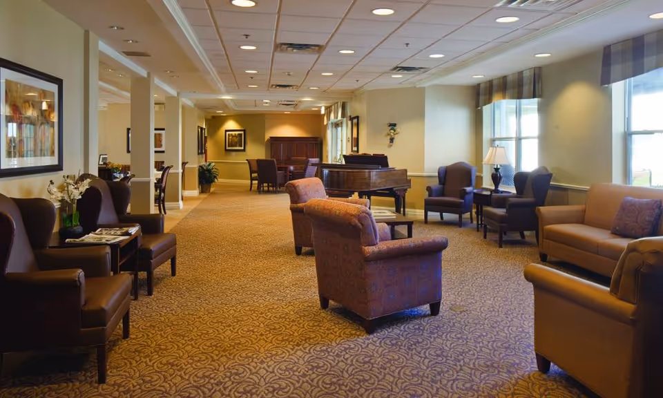 A spacious senior living common area with patterned carpet, multiple armchairs and sofas arranged for conversation, a grand piano, side tables with lamps, and large windows with valances allowing natural light to fill the room.
